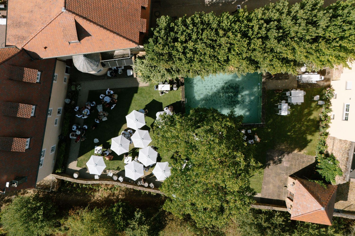 Aerial view of a charming outdoor restaurant courtyard with white umbrellas, pool, and lush green trees.
