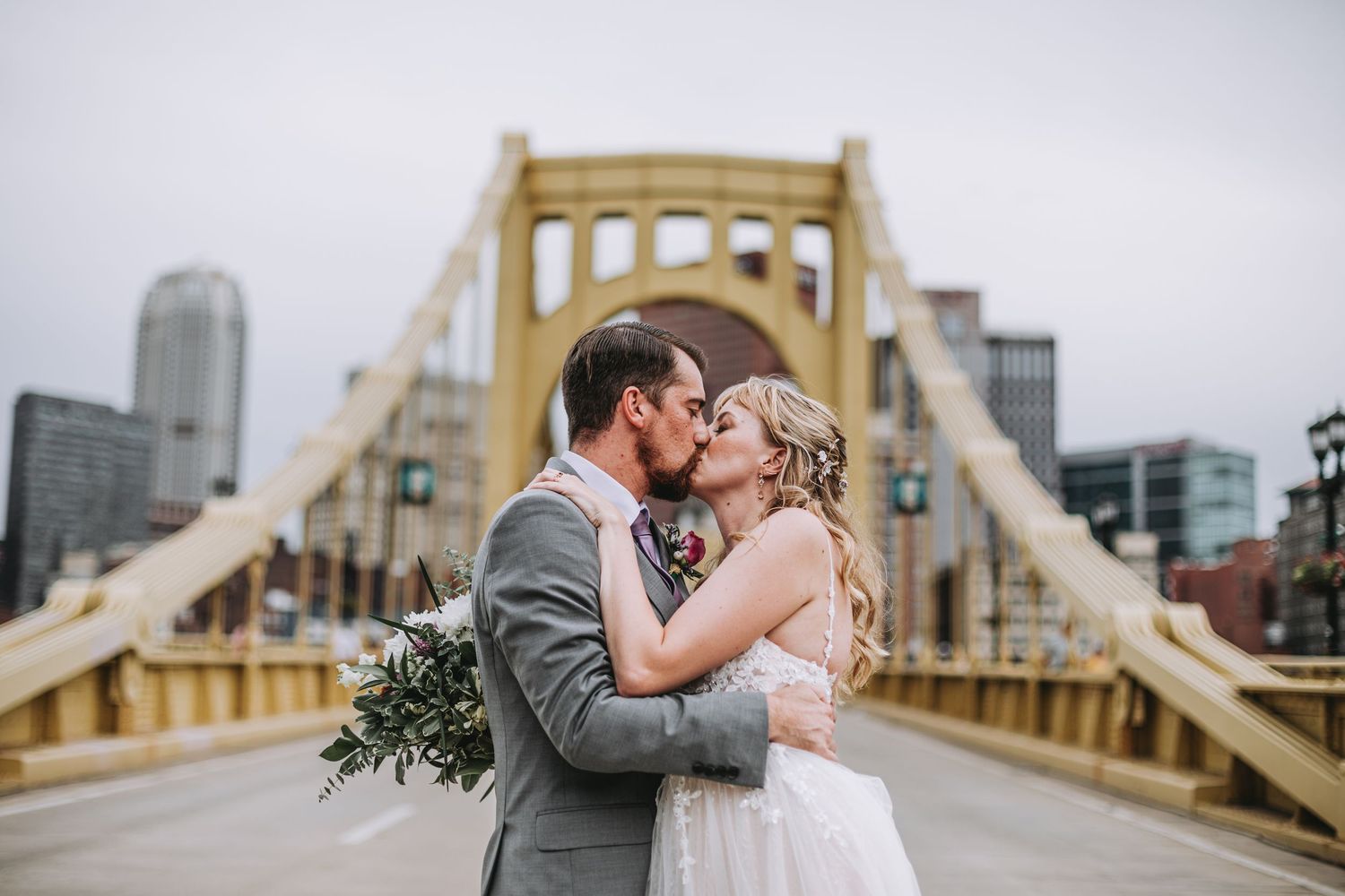Bride and groom share a romantic kiss on Pittsburgh's Roberto Clemente Bridge, with city skyline in the background.