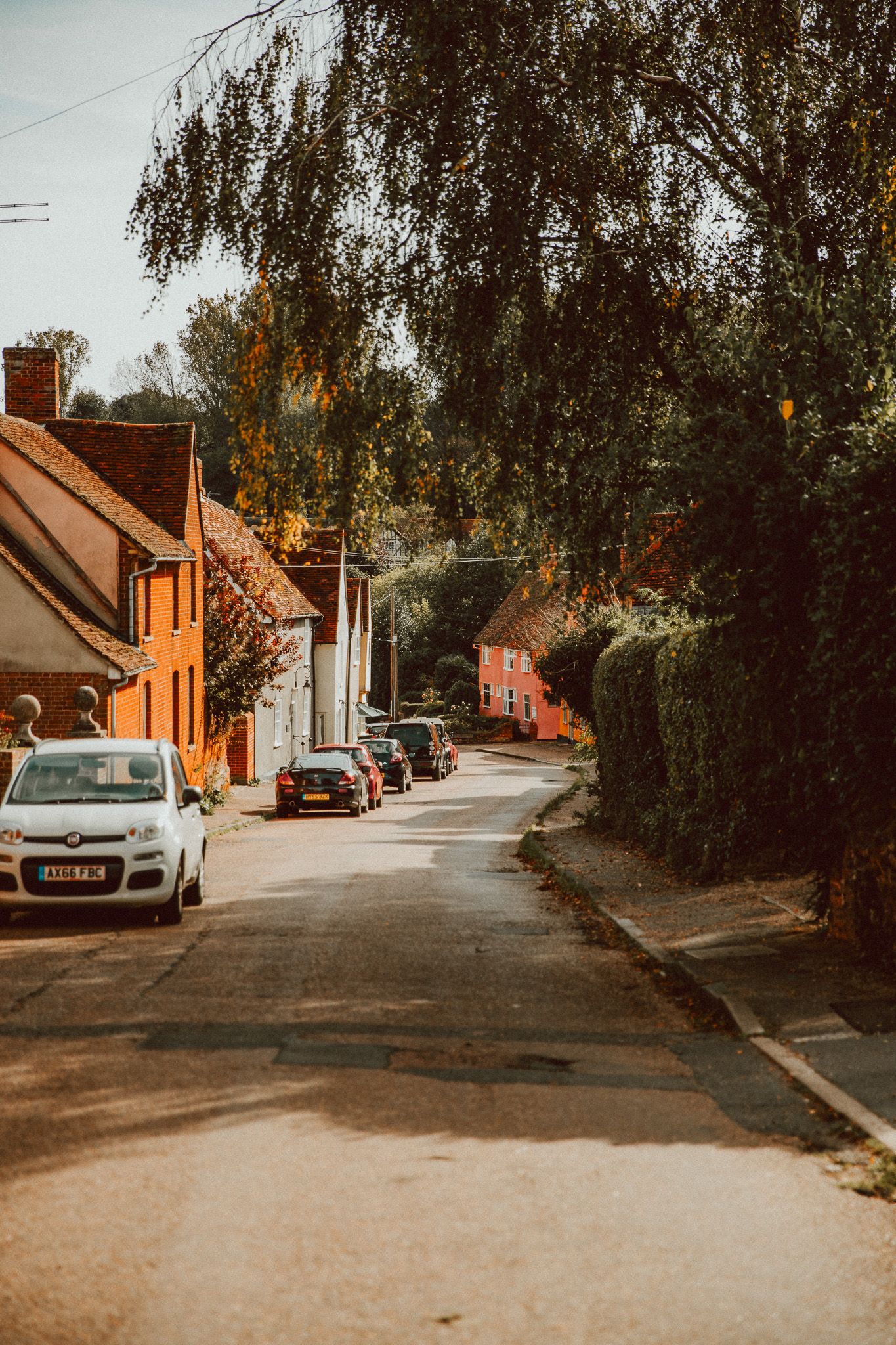 A quiet English village street lined with brick cottages, parked cars, and lush green trees on a sunny day.