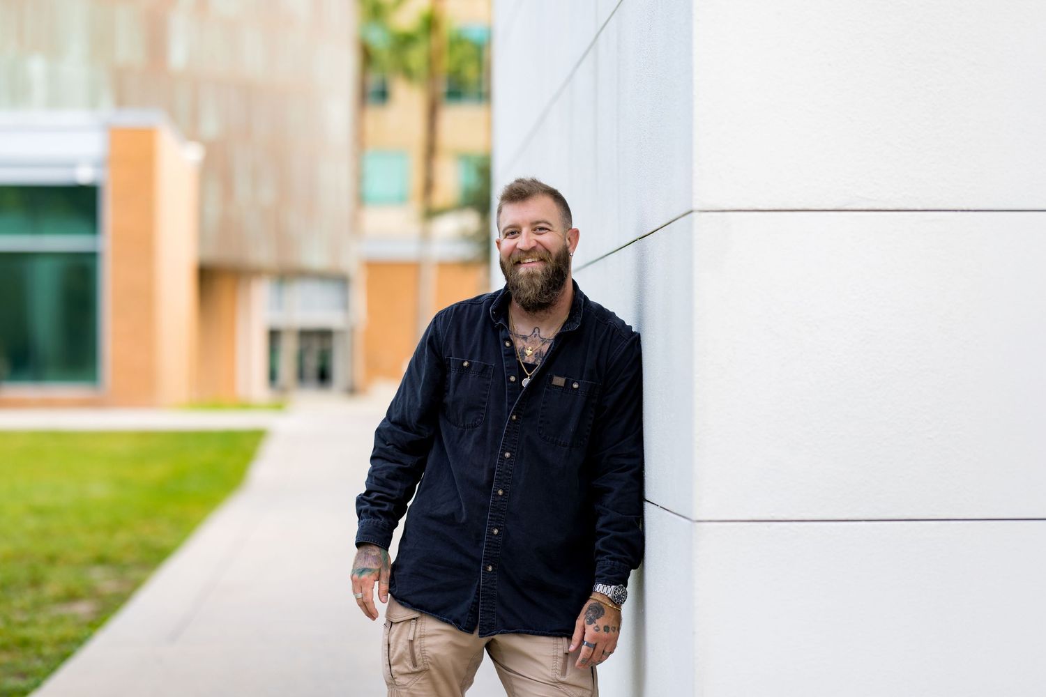Smiling bearded tattooed man in black shirt leaning against white wall outdoors near modern building.
