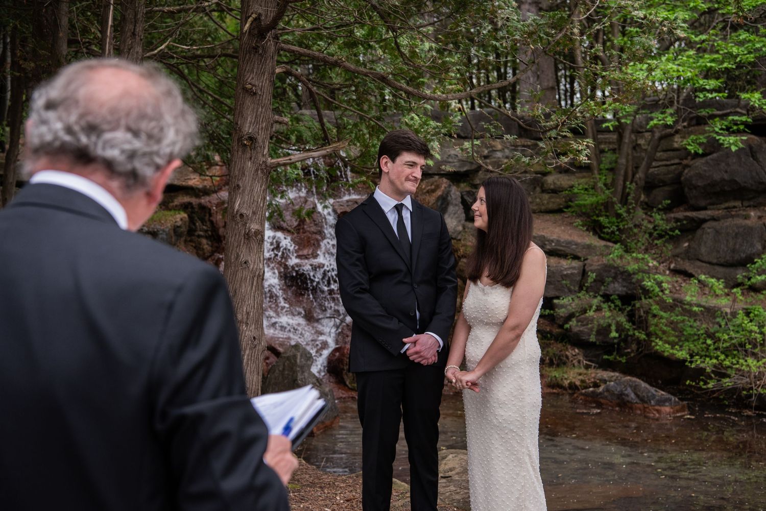 Couple stands before officiant during intimate forest wedding ceremony with cascading waterfall in background.