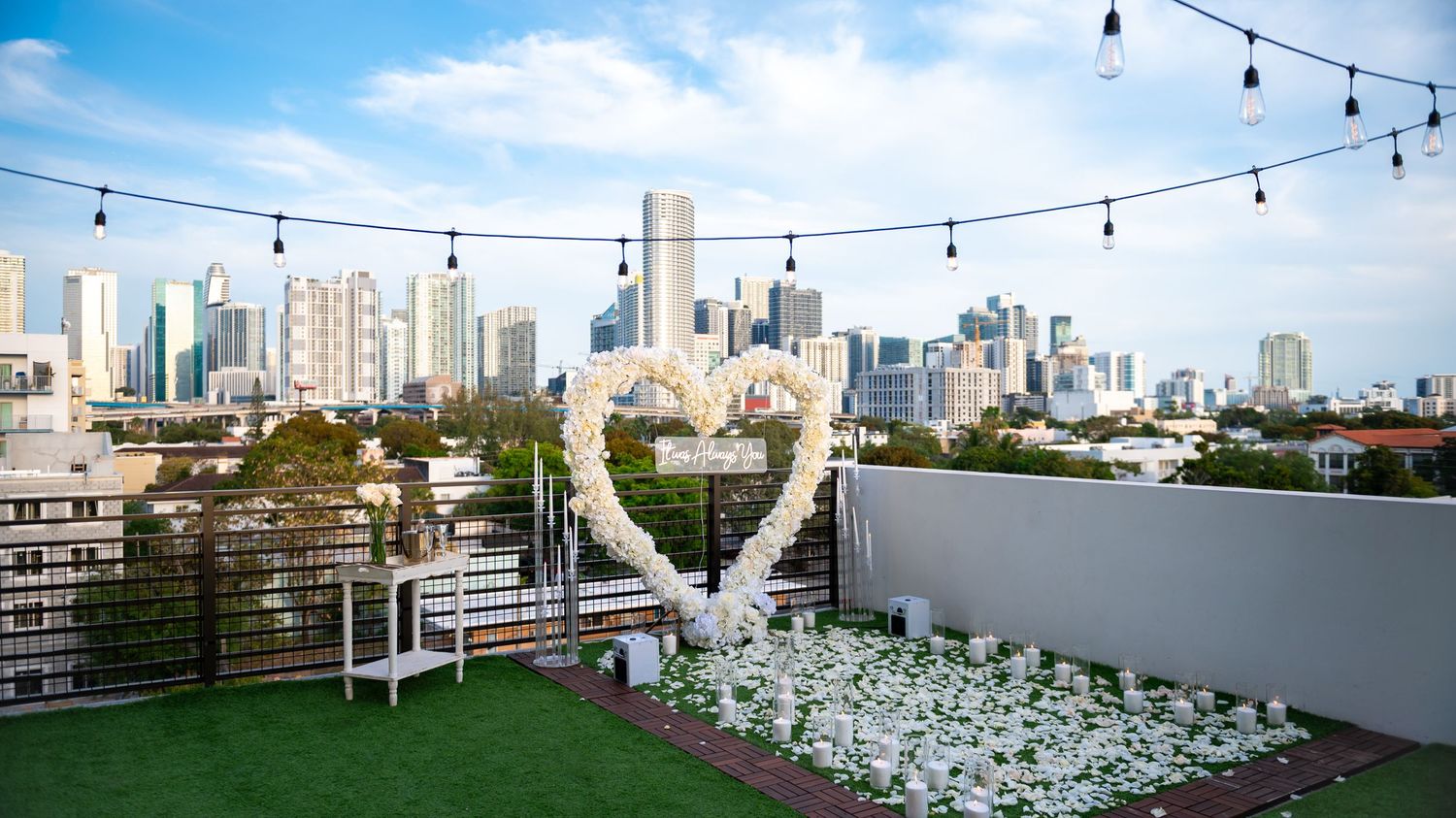 "Romantic rooftop proposal setup in Miami with rose petals, candles, and an 'It Was Always You', overlooking the city skyline at sunset."