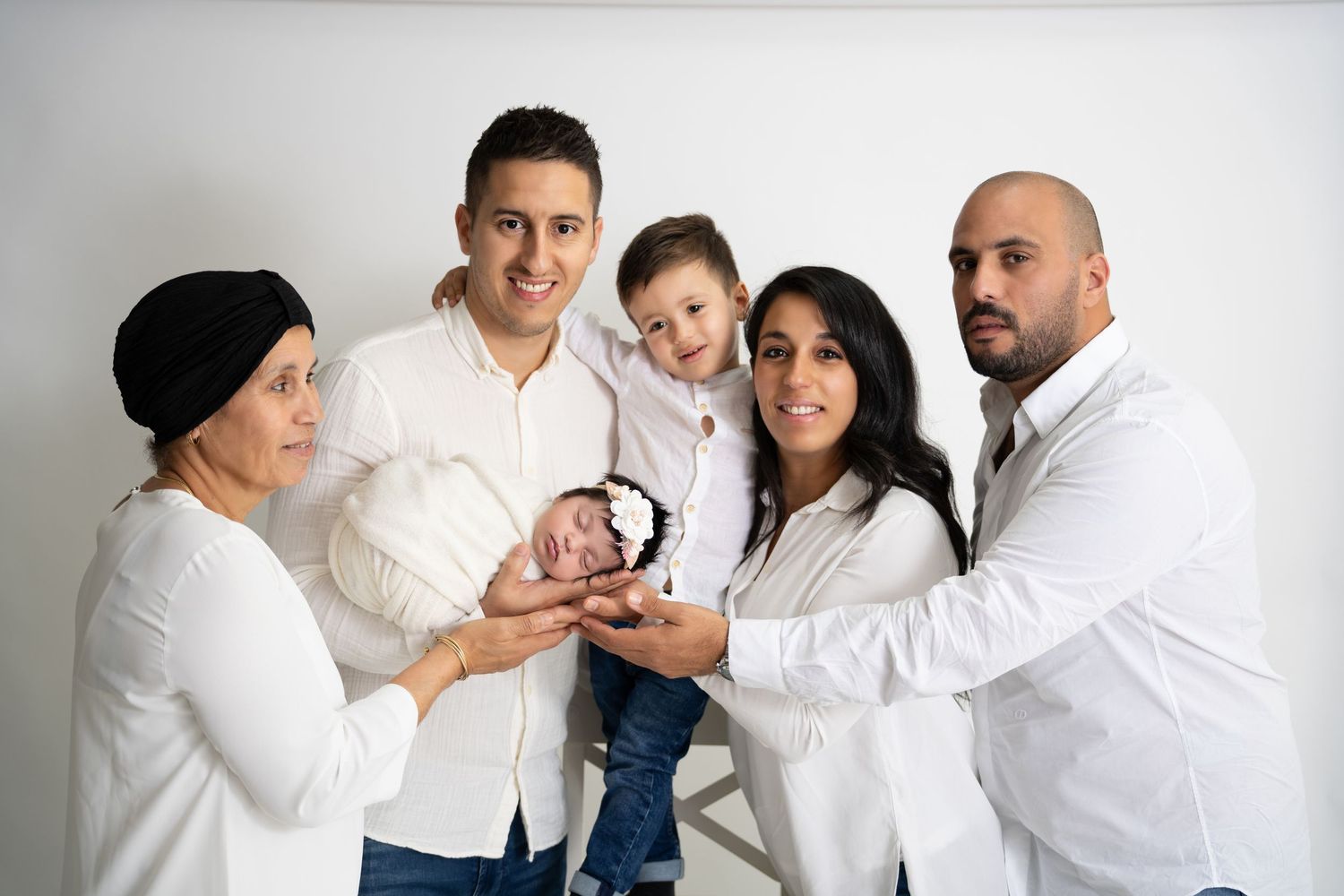 A family in white attire holding their newborn baby together against a light gray studio background.