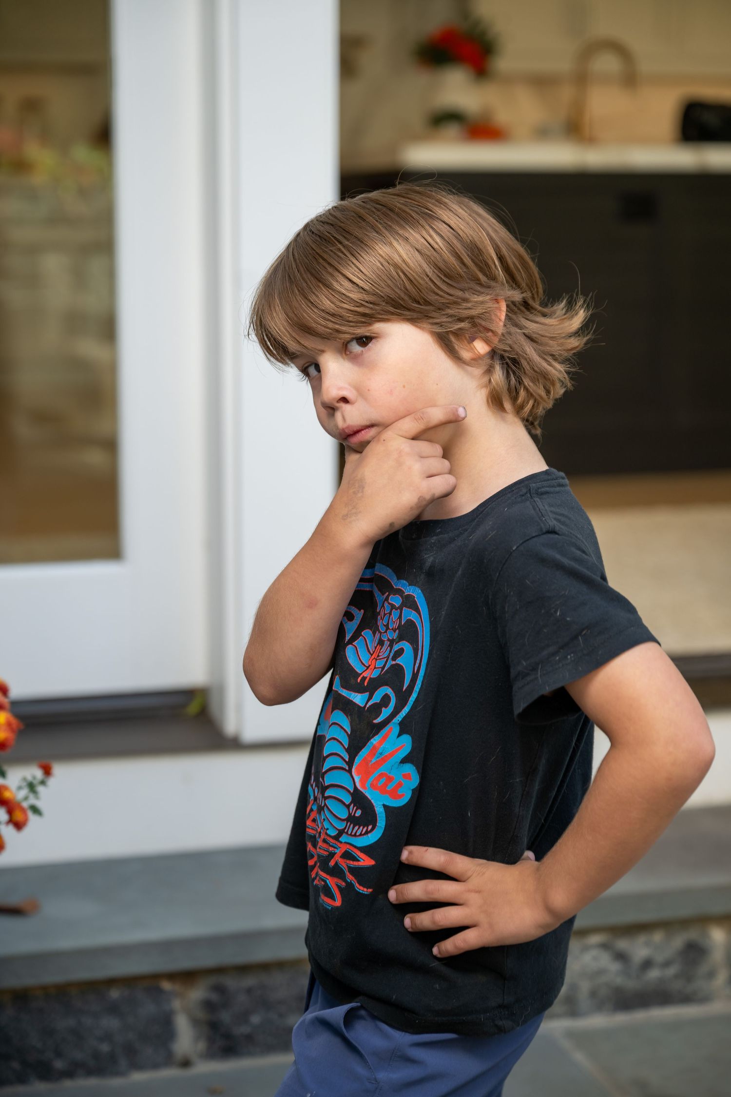 Child wearing a dark t-shirt with blue graphic design stands in thoughtful pose against blurred kitchen background.