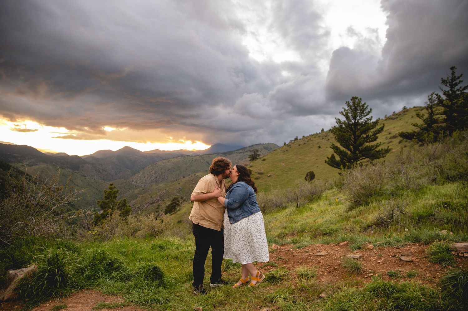 A couple embraces on a mountain ridge overlooking valleys and hills under dramatic stormy sunset skies with evergreen trees.