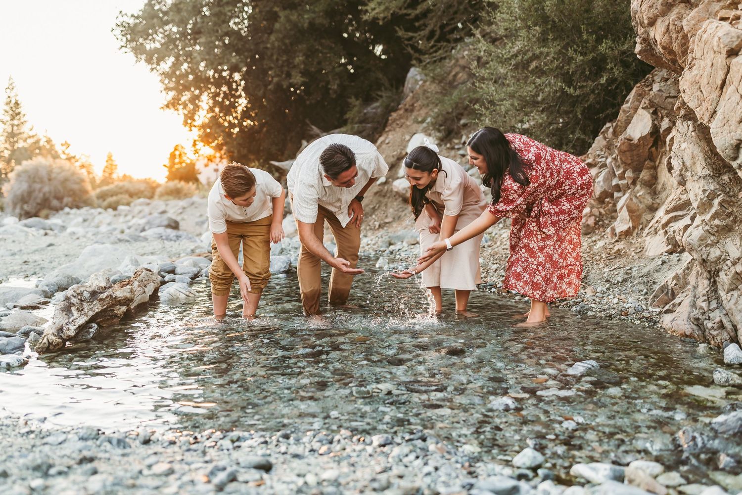 A group in light summer clothing bends down to explore a rocky shoreline at sunset.