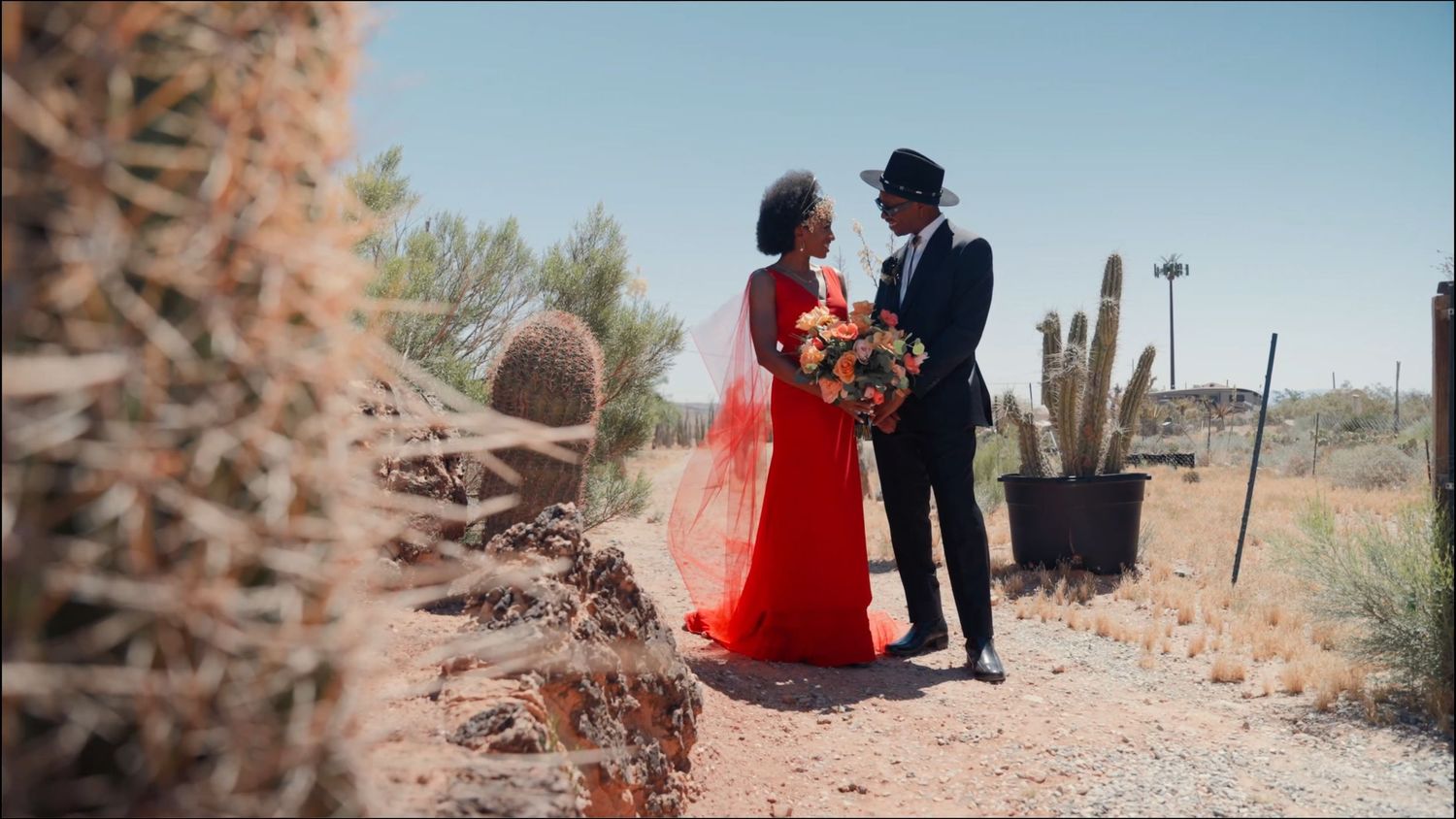Couple in red dress and black suit exchanging vows in desert landscape with cacti.