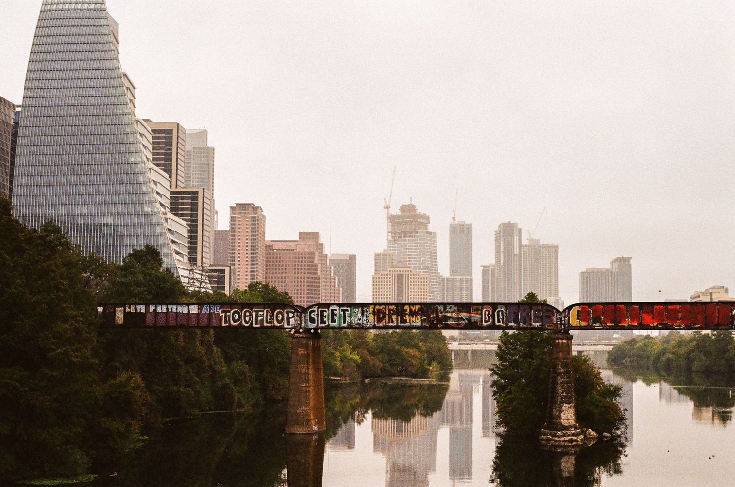 Urban skyline with railroad bridge crossing reflective river under overcast sky.
