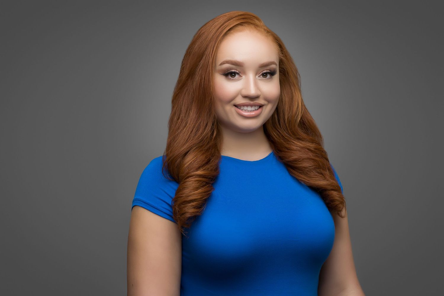 Professional portrait of person in blue shirt with red hair against gray background.