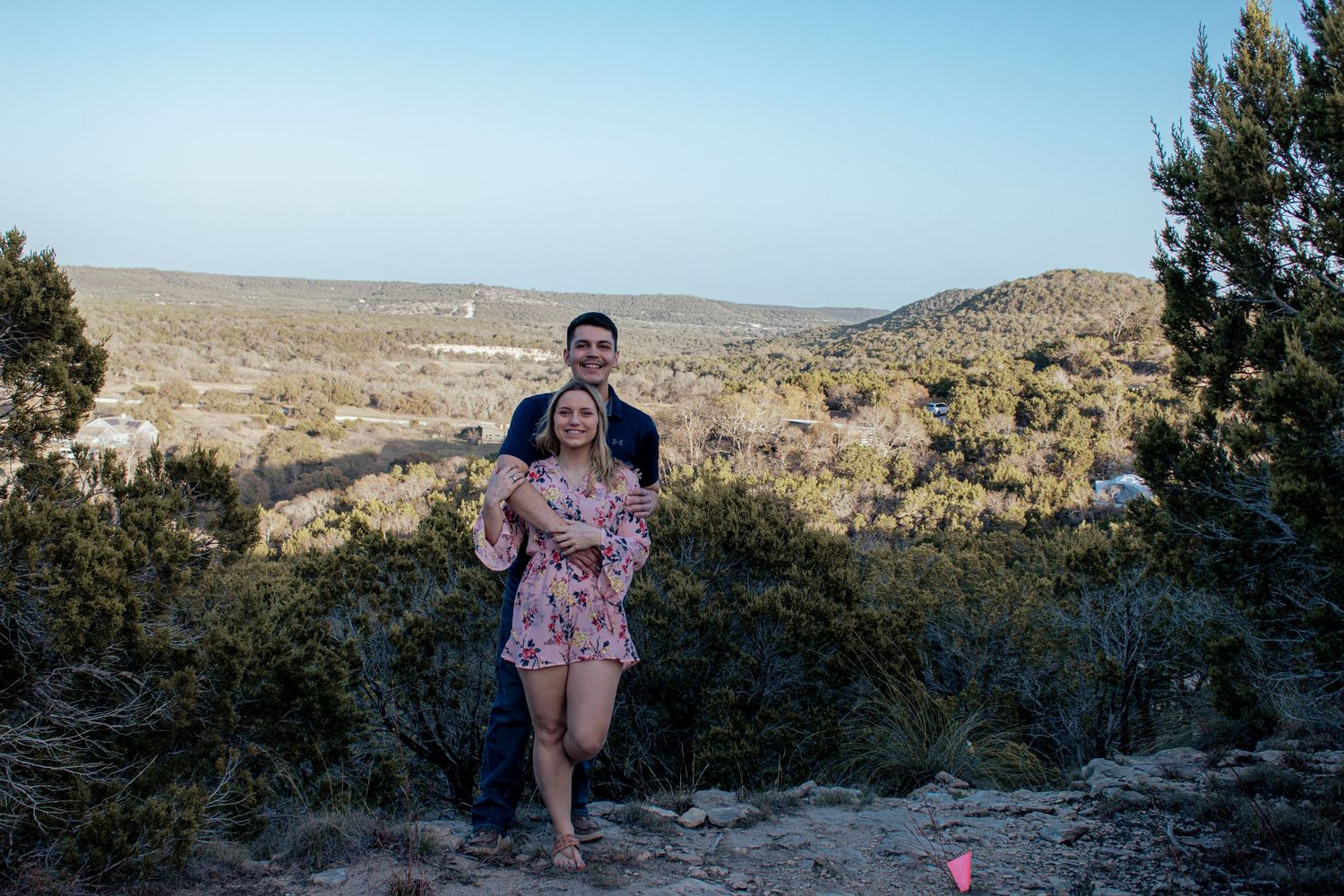 A couple stands on a rocky overlook with desert hills and mountains stretching into the distance.