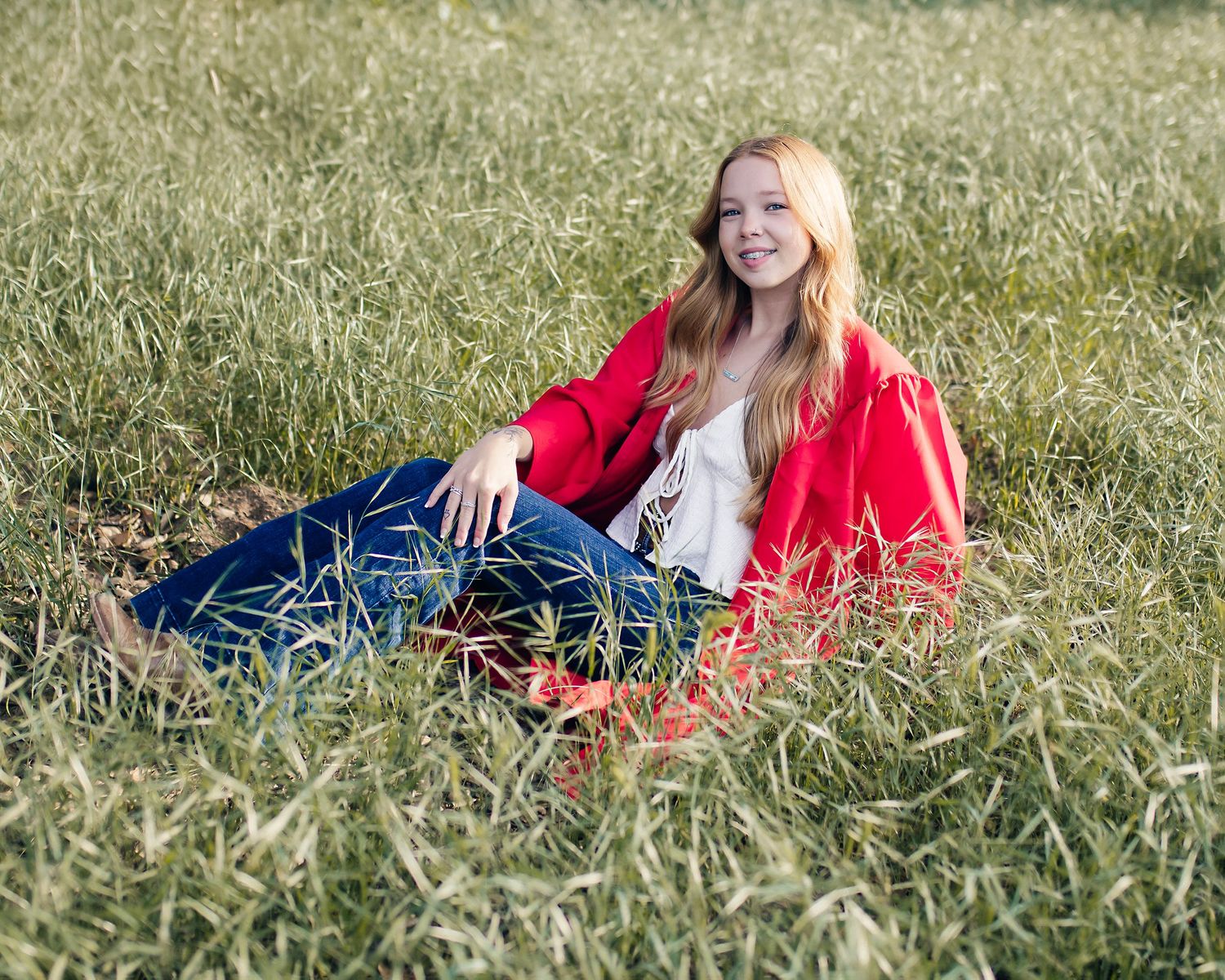 Graduate relaxing in grass wearing red graduation gown and casual denim.