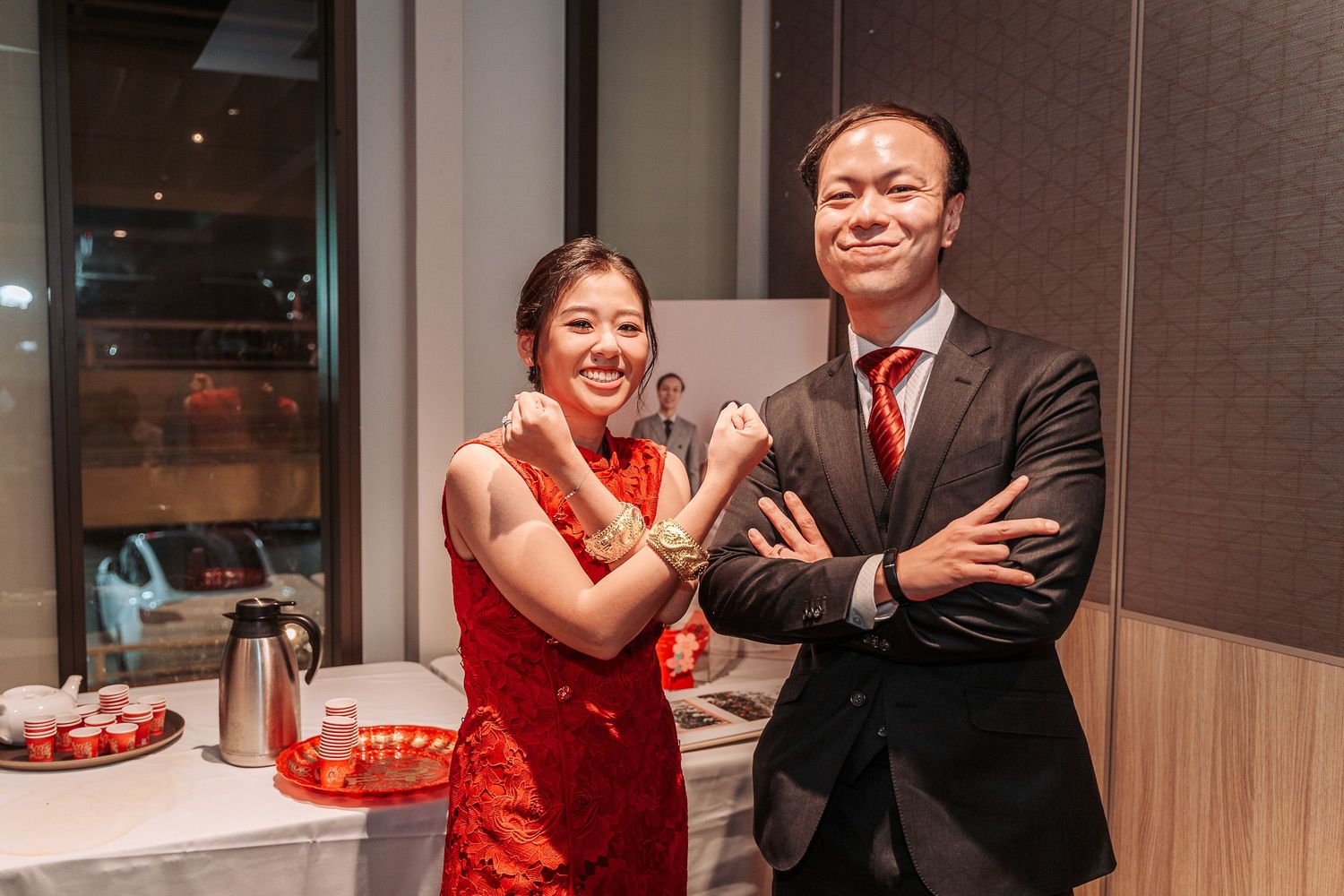 Two people at a festive celebration wearing traditional Chinese red dress and black suit stand together smiling indoors.