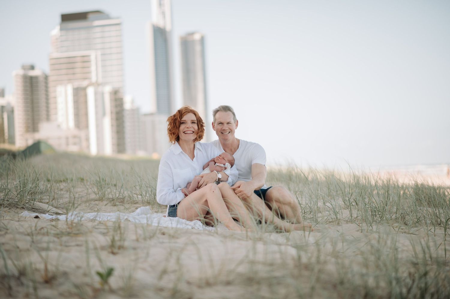 Couple sitting on coastal dunes with city skyline visible in the background.