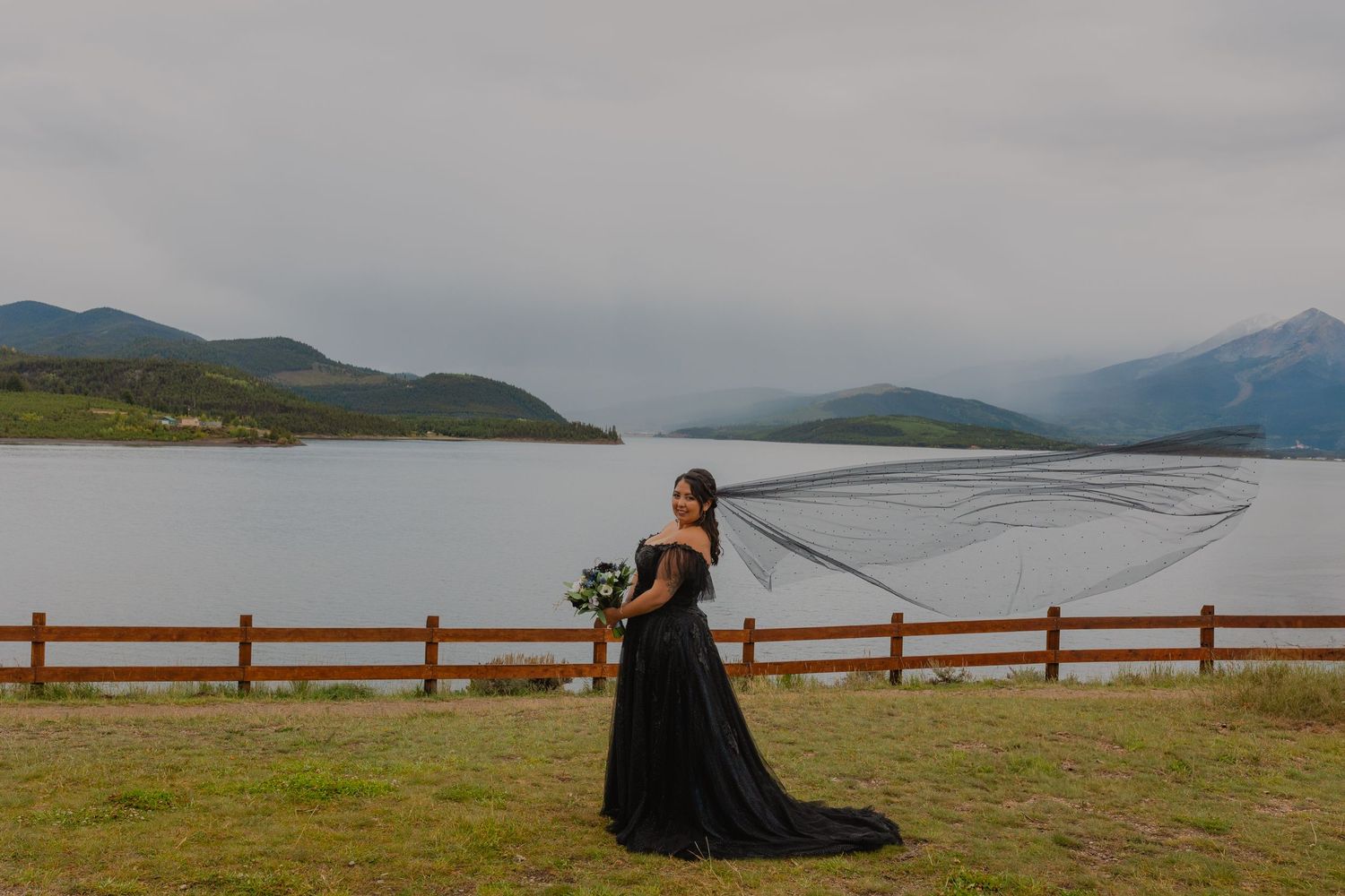 Someone in a black flowing dress stands near a wooden fence overlooking a mountain lake on a cloudy day.