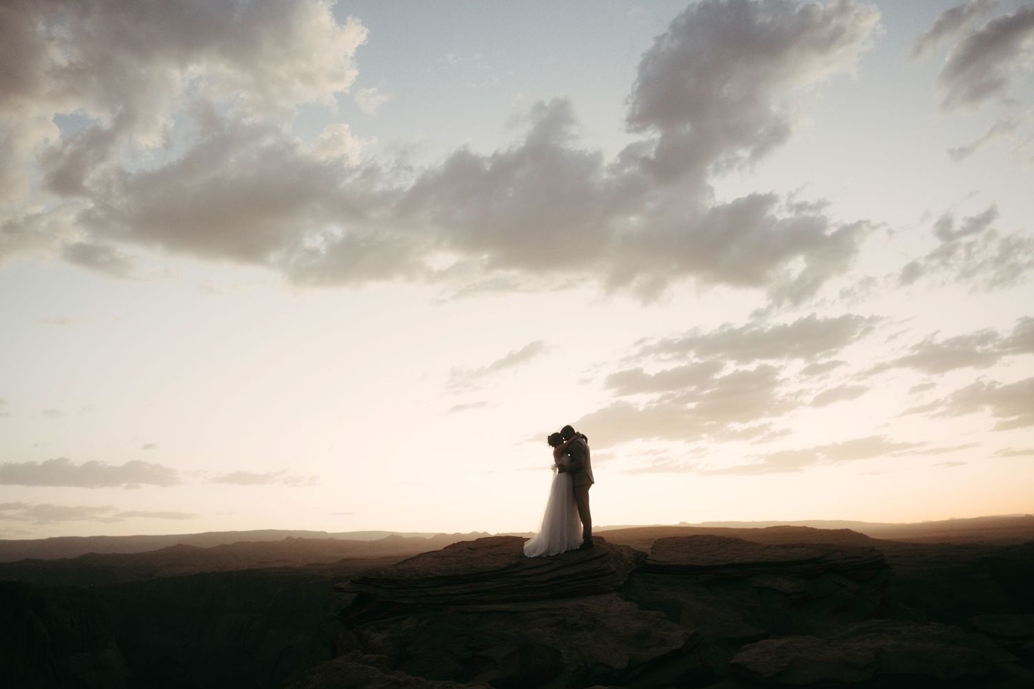 Silhouetted couple stands together on a hilltop against a dramatic sunset sky with wispy clouds.