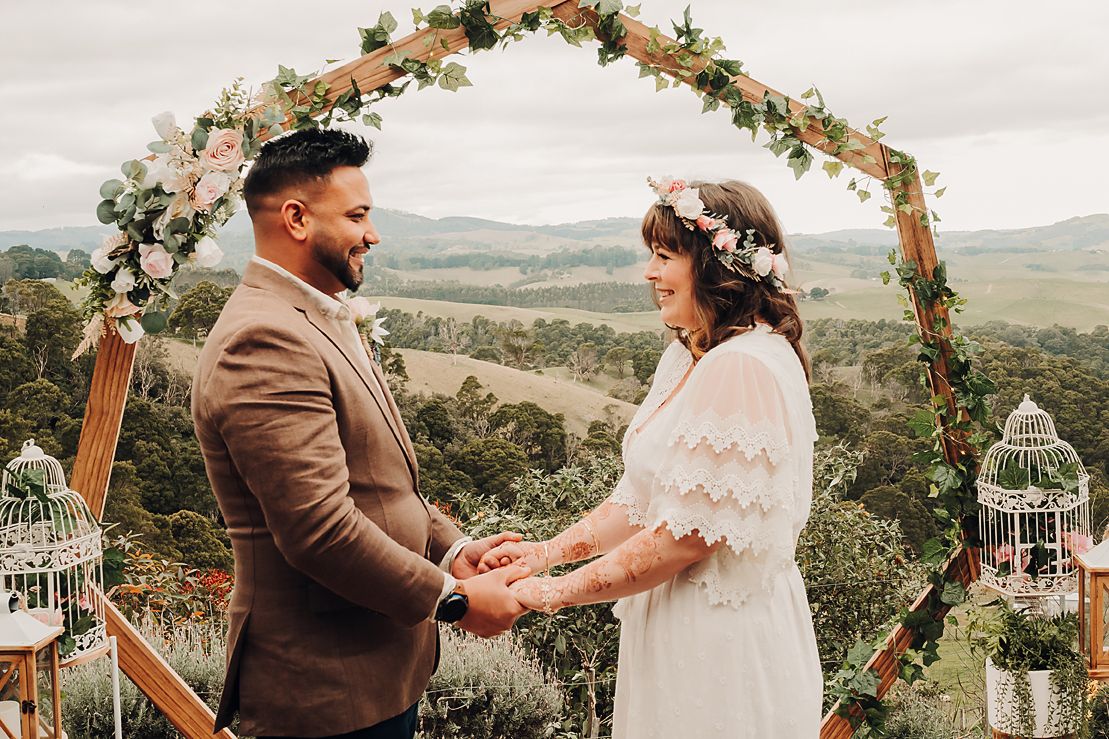 A romantic outdoor wedding ceremony under a floral arch with rolling hills in the background.