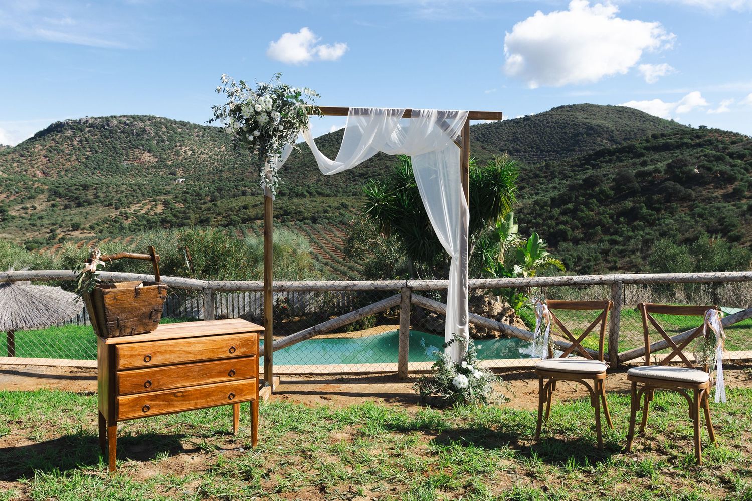 Rustic outdoor ceremony setup with wooden bench, arch with white drapes, and mountain views in the background.