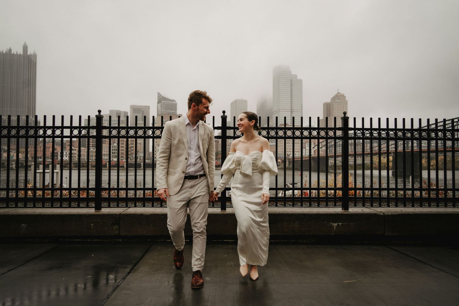 A couple in light-colored attire walks together on a rainy city rooftop with a misty skyline in the background.