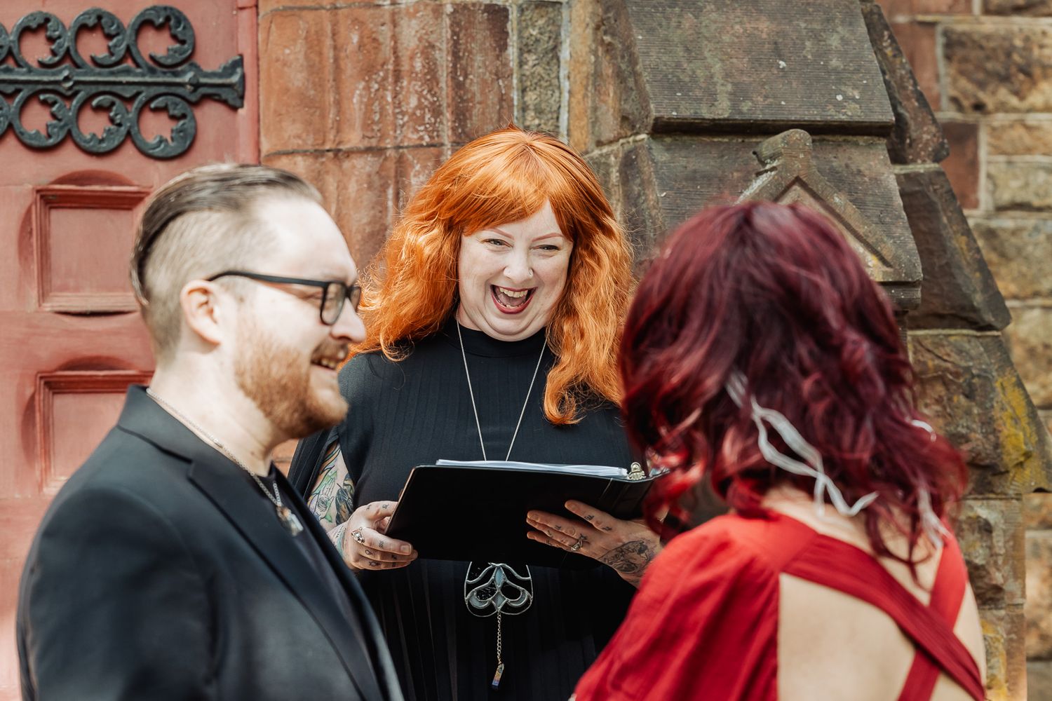Three people share laughter during outdoor ceremony against stone building backdrop.