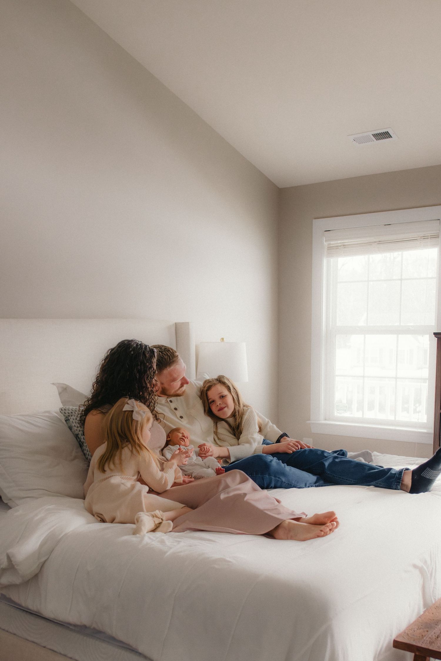 A family relaxes together on a cozy white bed near a bright window in a minimalist bedroom.