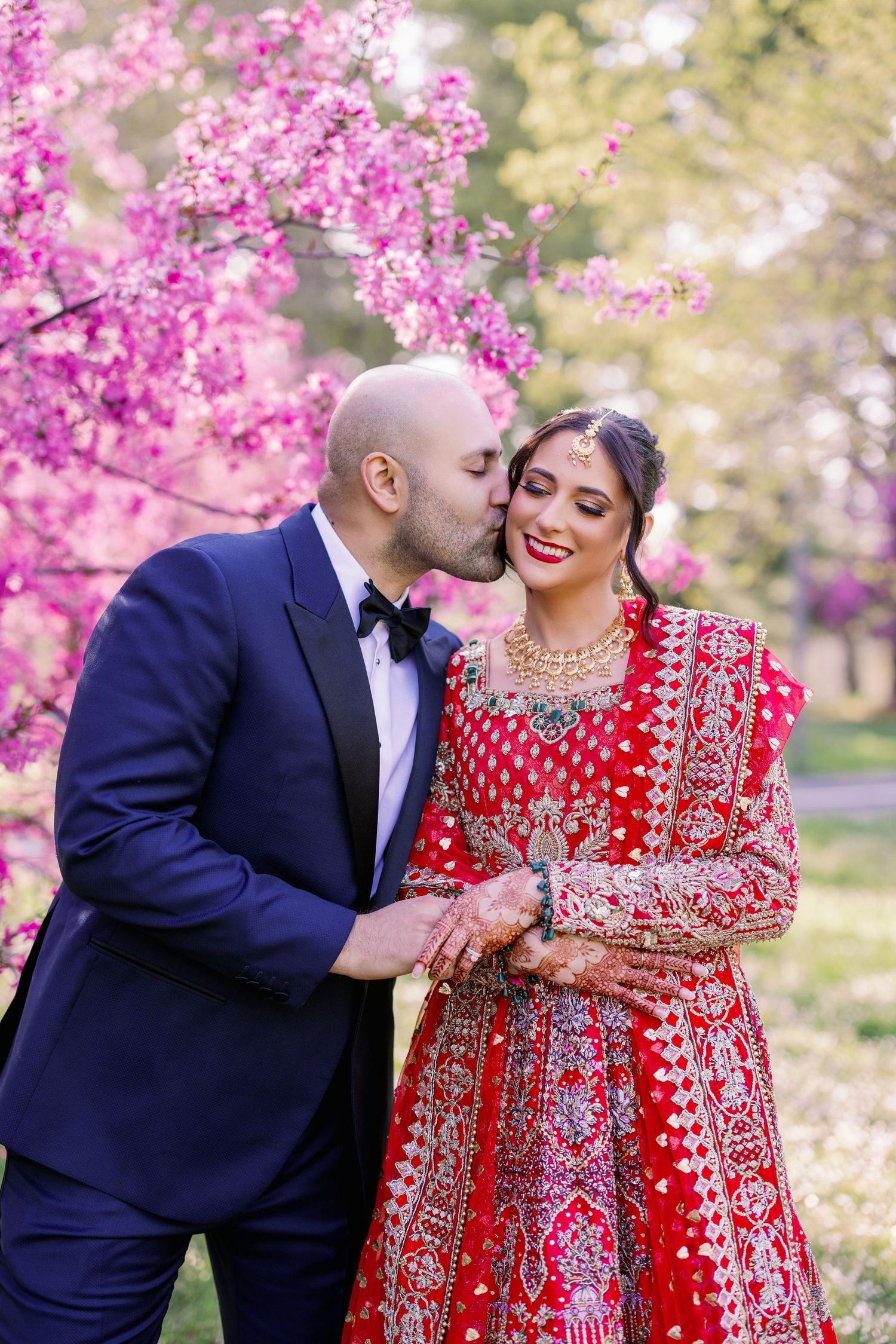 A couple shares a romantic moment under blooming cherry blossoms, with the bride wearing a traditional red lehenga.