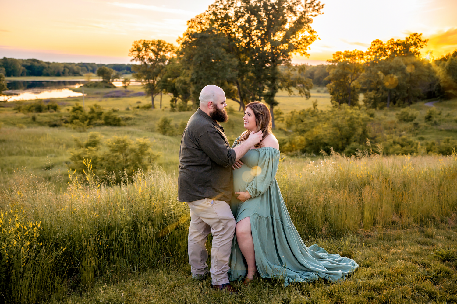A couple shares a romantic moment in a golden sunset field by a lake with trees silhouetted in the background.