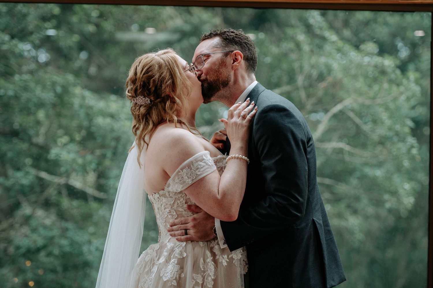 A romantic outdoor wedding moment with a couple sharing a kiss against a lush green forest backdrop.
