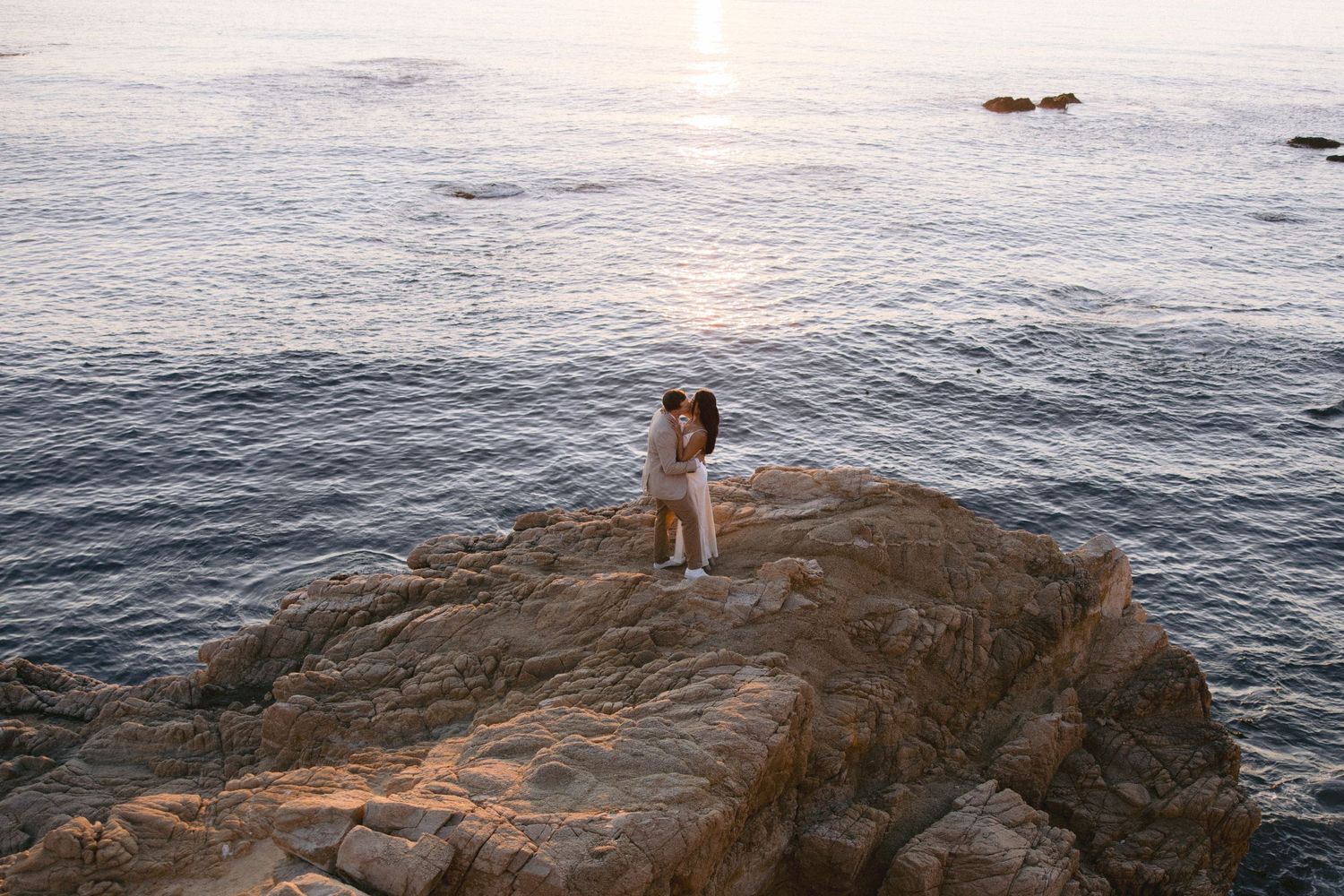 An embracing couple stands on a rocky ocean cliff during a serene sunset over calm waters.