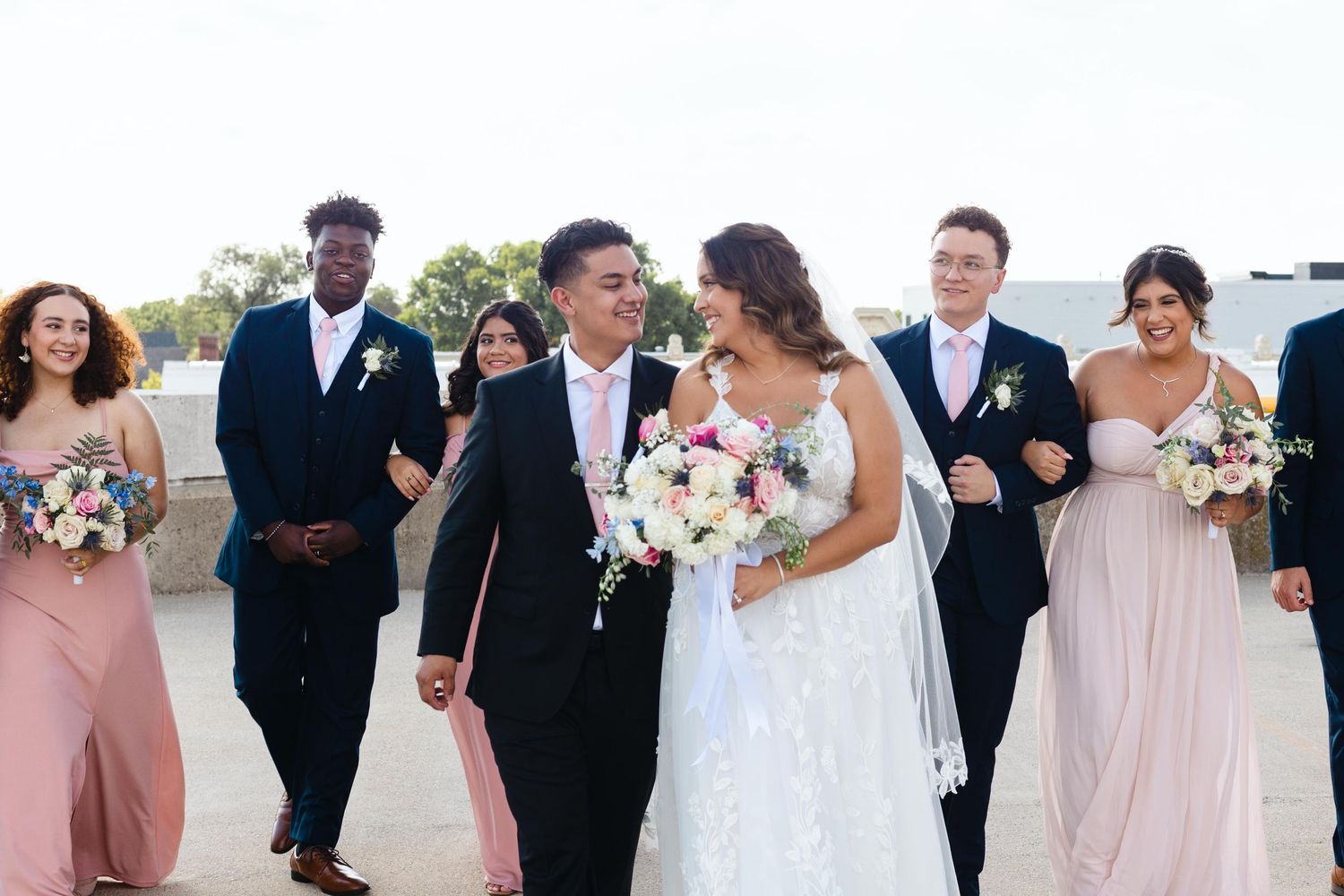 Wedding party walking together outdoors with bride in white gown and bridesmaids in blush pink dresses holding bouquets.