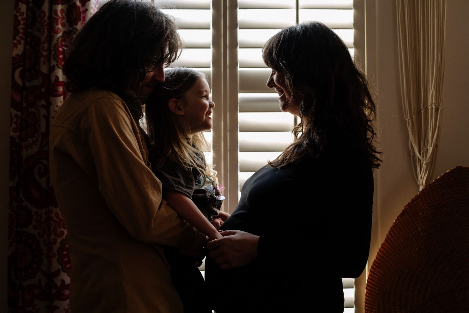 Three silhouetted figures stand together by a window with curtains and shutters casting shadows in soft natural light.