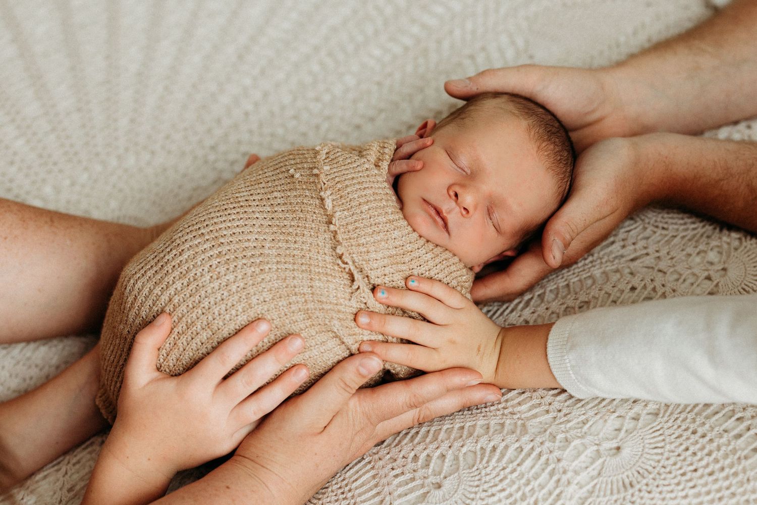 Newborn baby wrapped in knit sweater lying on white blanket with hands gently touching.