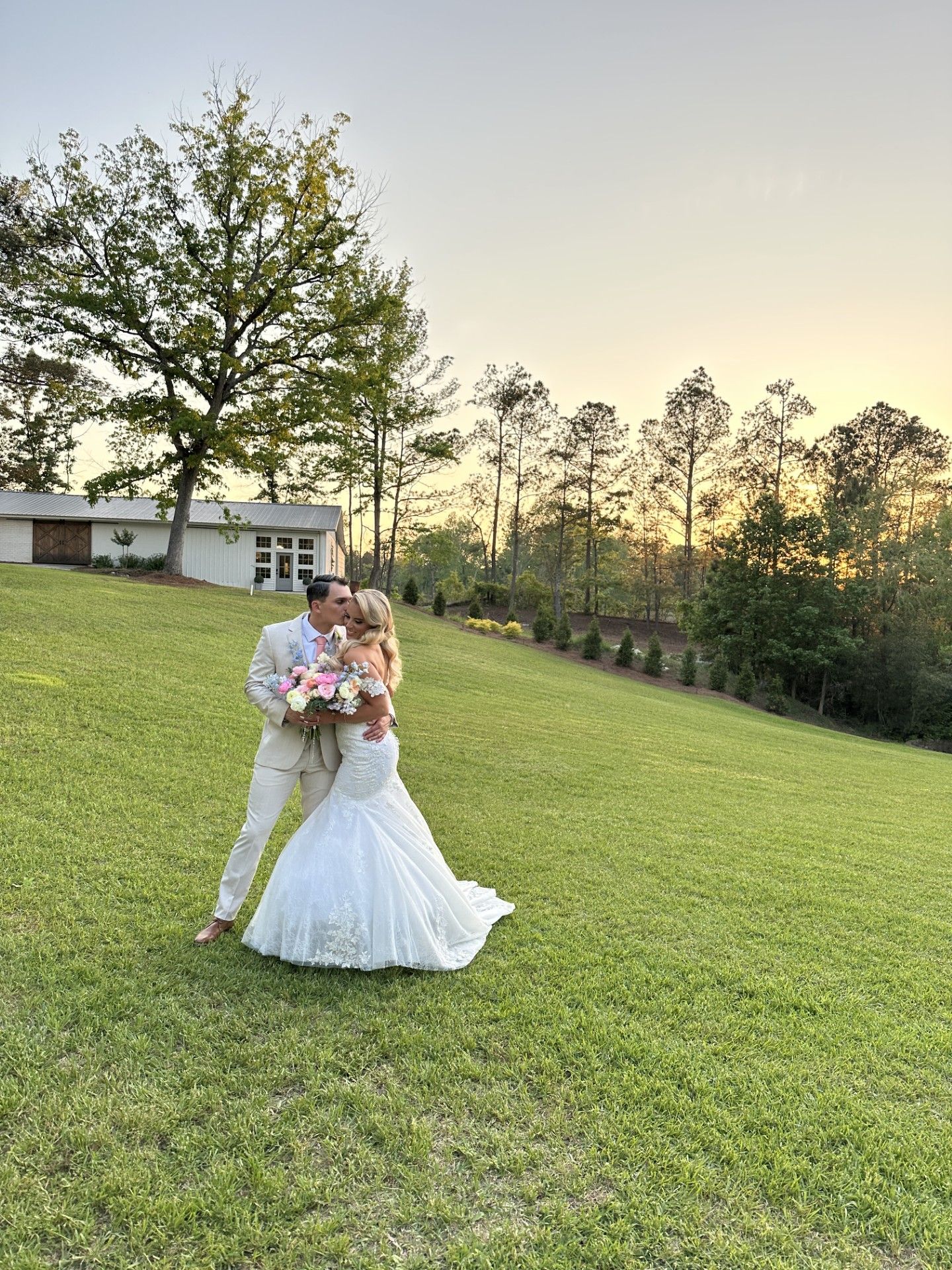 A couple shares a romantic moment on a grassy hill at sunset with trees and a building in the background.