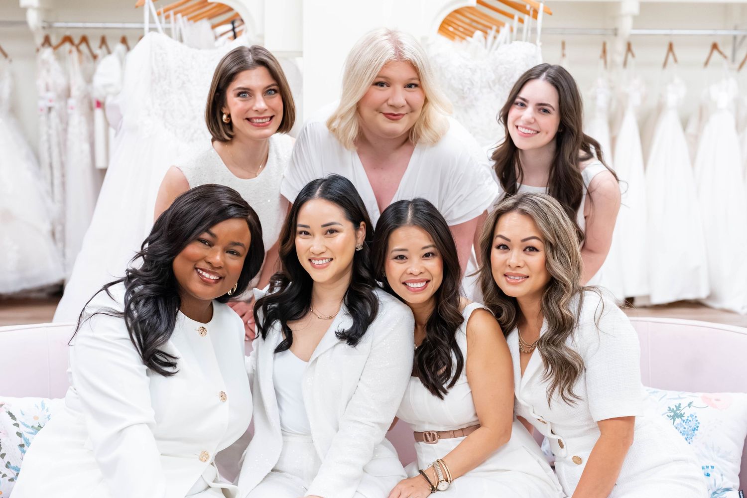 Seven women in white outfits smiling together inside a bridal boutique with wedding dresses displayed in the background.