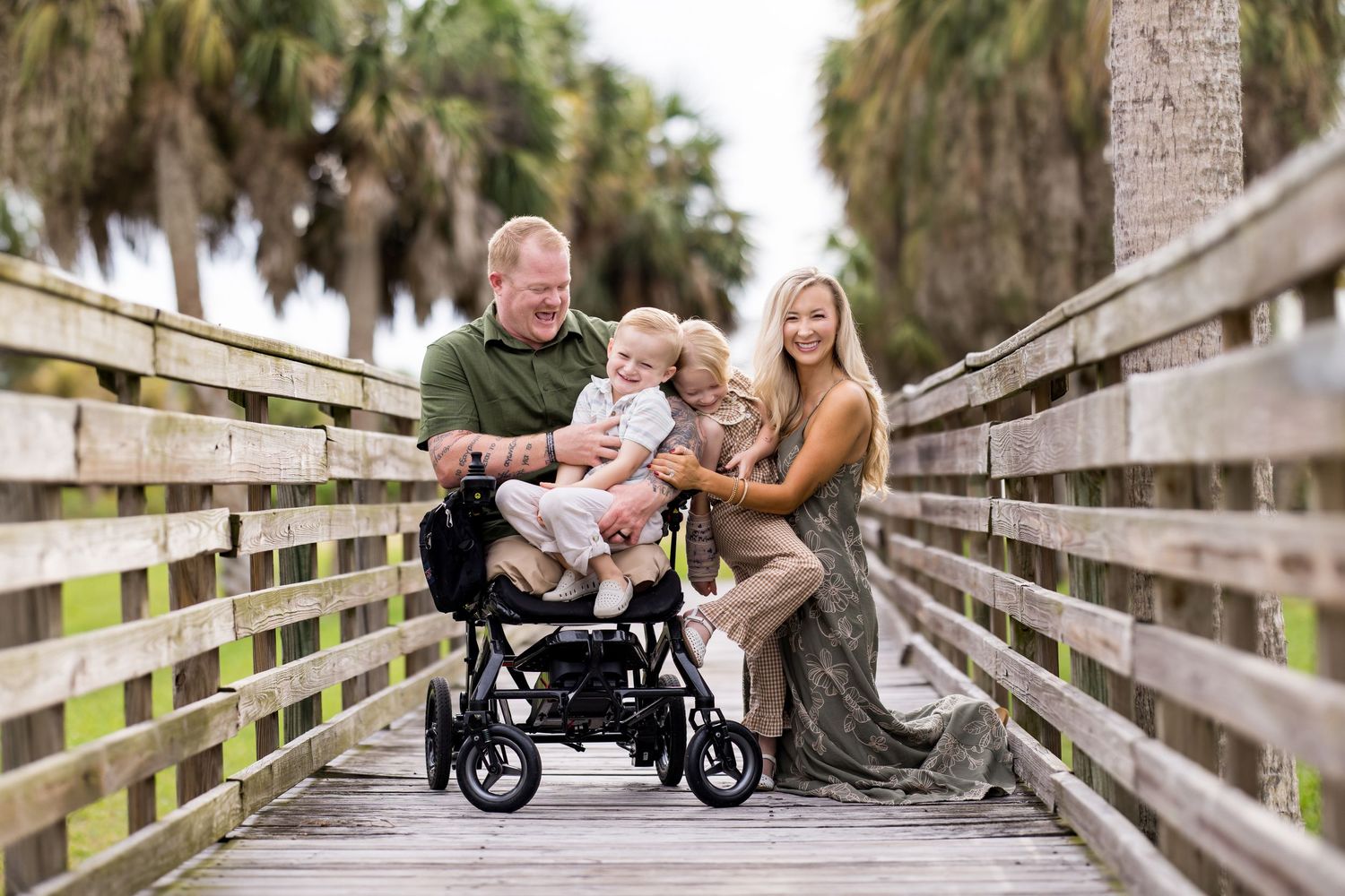 Family smiling on a wooden boardwalk surrounded by palm trees, with a young child in a stroller.