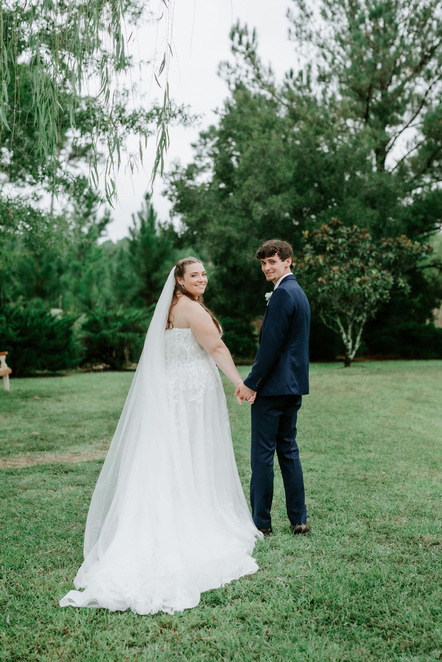 Bride and groom walk hand-in-hand under a wooden pergola, she holds blue floral bouquet, guests visible behind them.