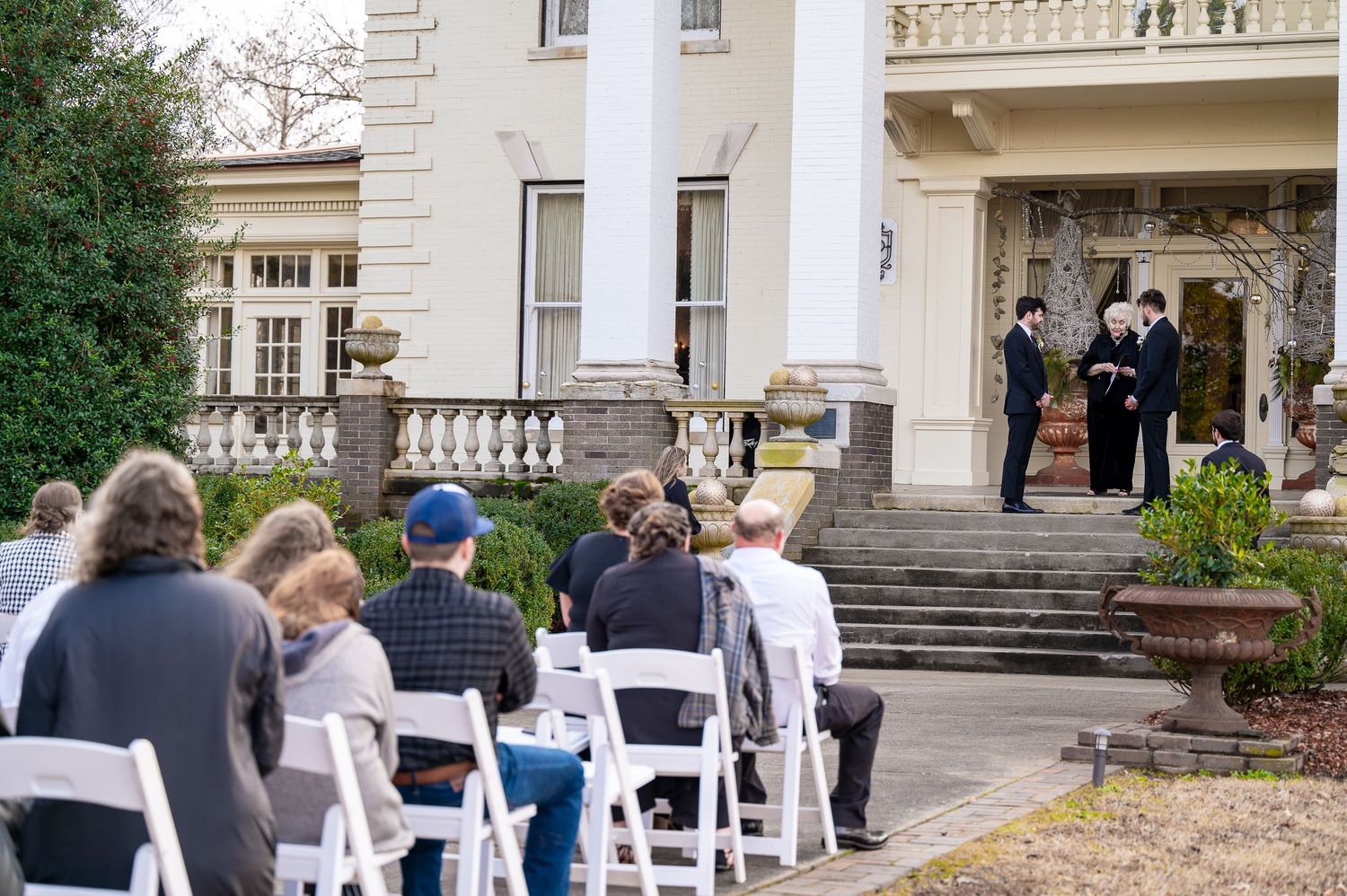 Outdoor wedding ceremony at elegant white mansion with guests seated in white chairs on manicured grounds.