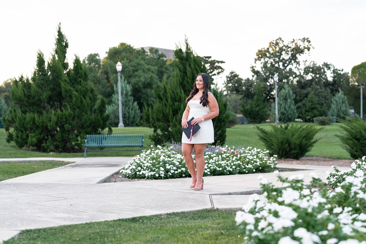 Woman with long dark hair in a white dress stands barefoot on a park path beside white flowers, evergreen trees and a green bench.