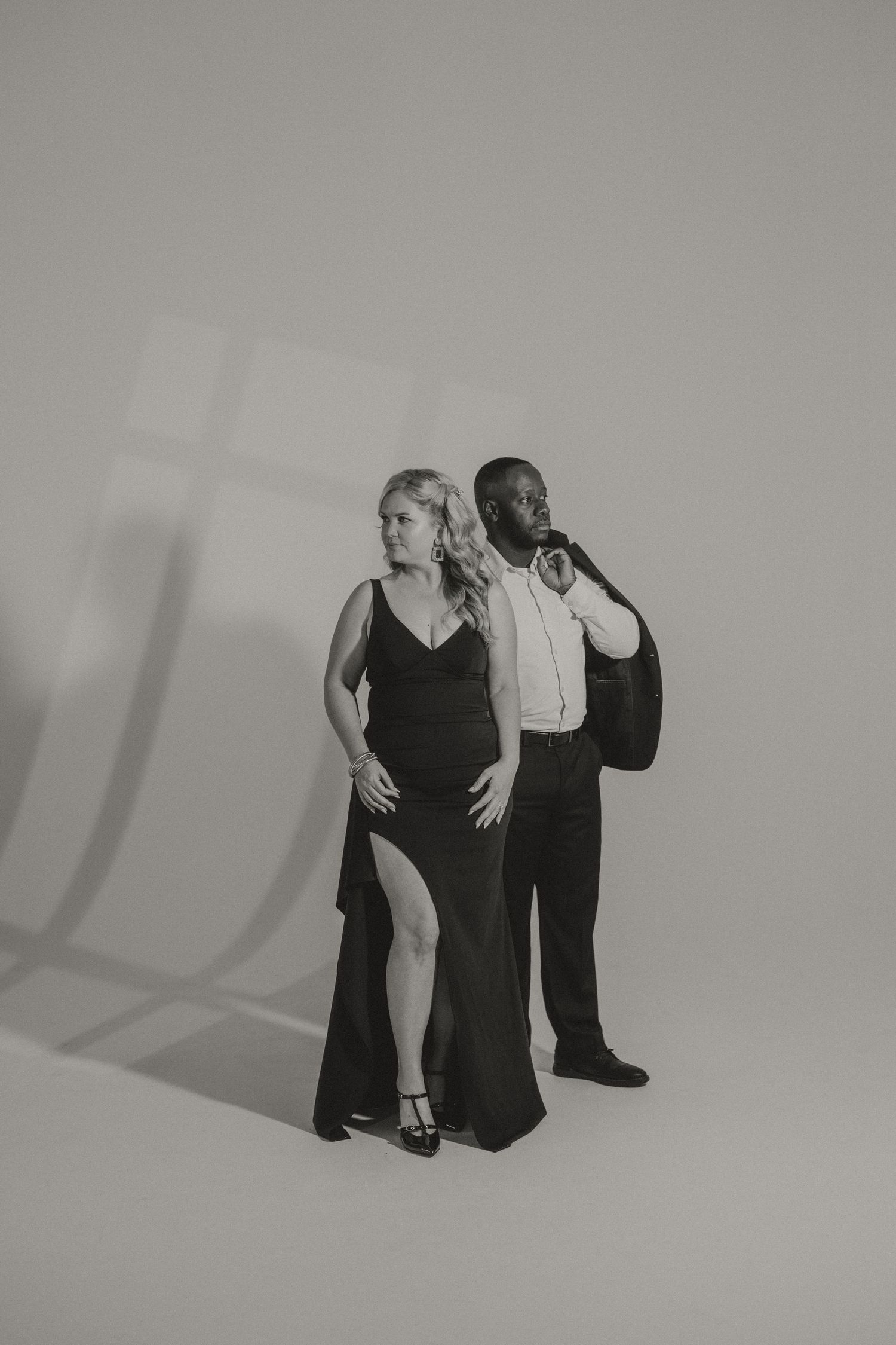 Black and white studio portrait of an elegant couple, the woman in a long slit gown and the man in a suit with his jacket over his shoulder.