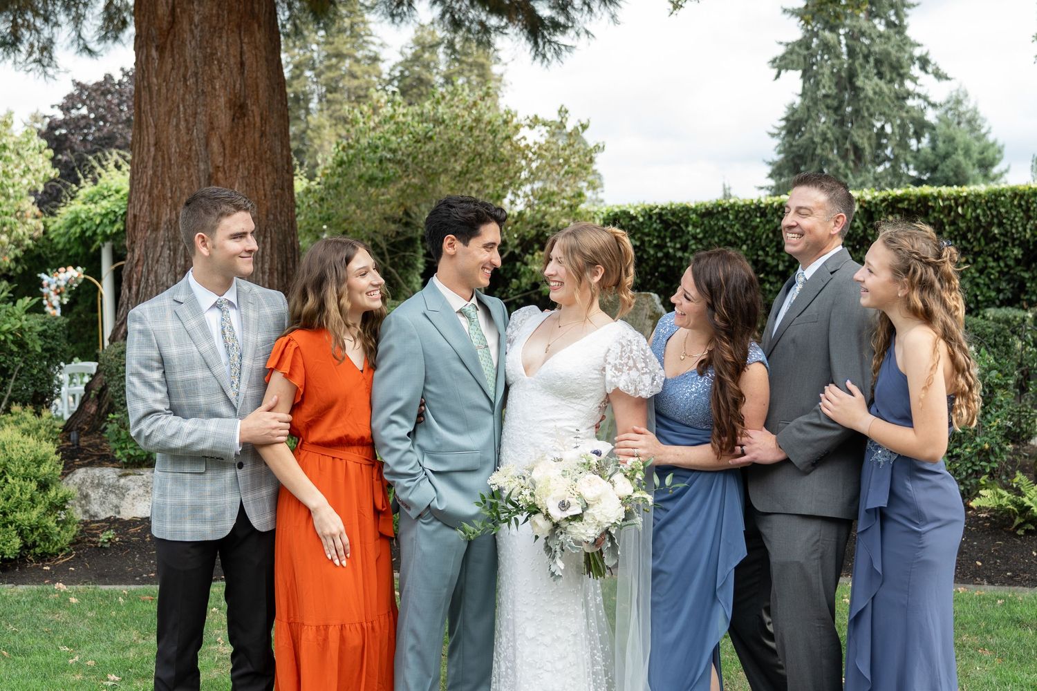Wedding party in garden, bride in white gown holding bouquet, groom in blue suit, guests in blue and orange dresses.