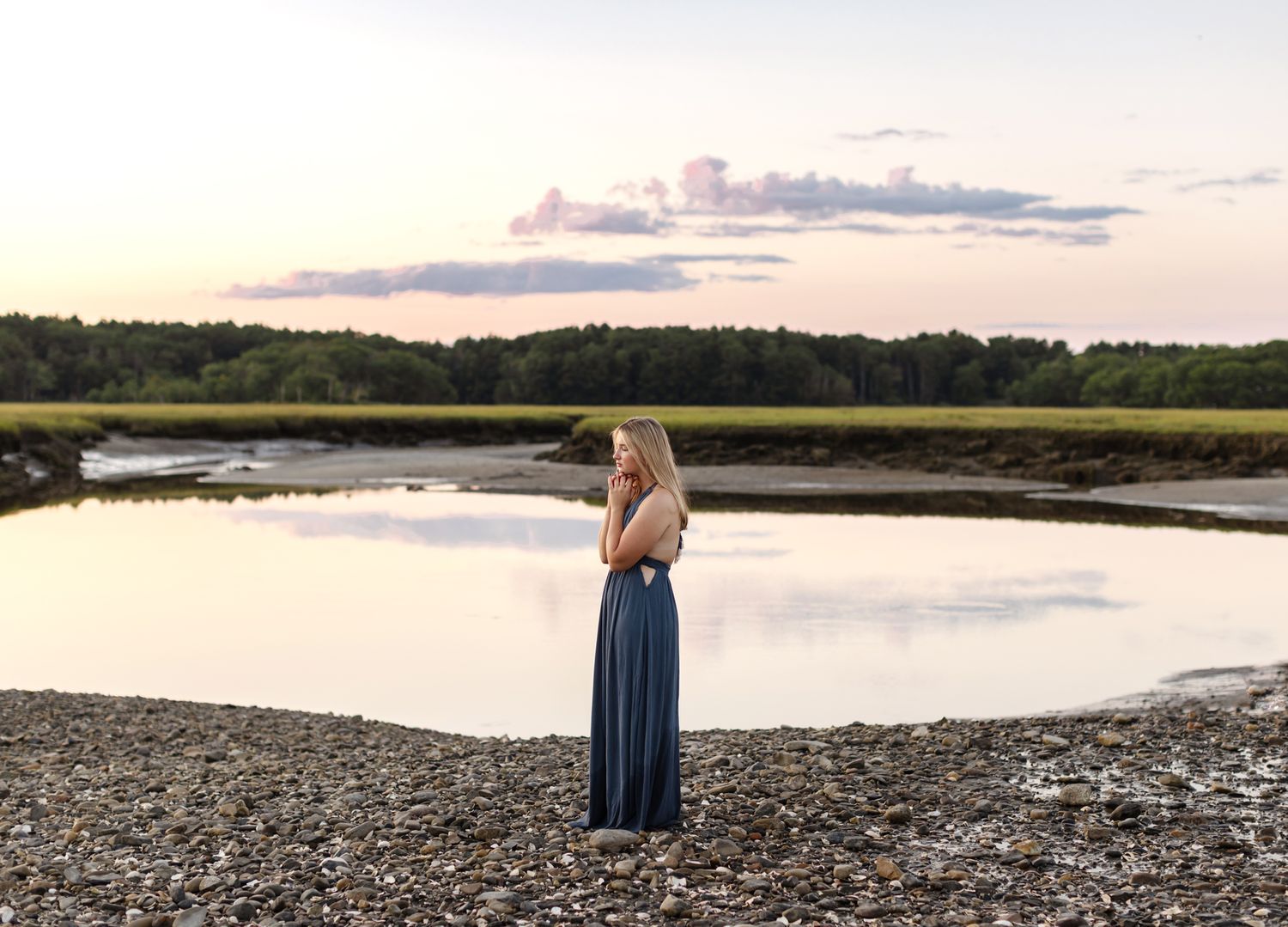 Young lady in blue maxi dress stands on rocky grassy cover at sunset with calm reflective water and trees in background for senior photos.