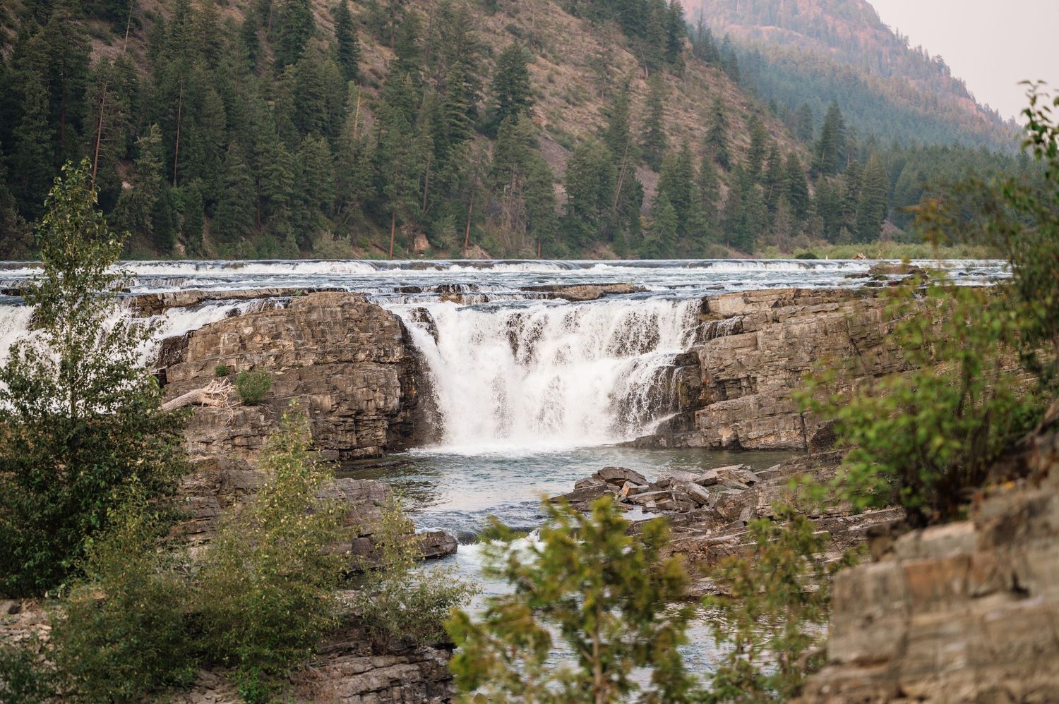 A scenic waterfall cascades over rocky ledges surrounded by evergreen trees in a misty mountain river landscape.