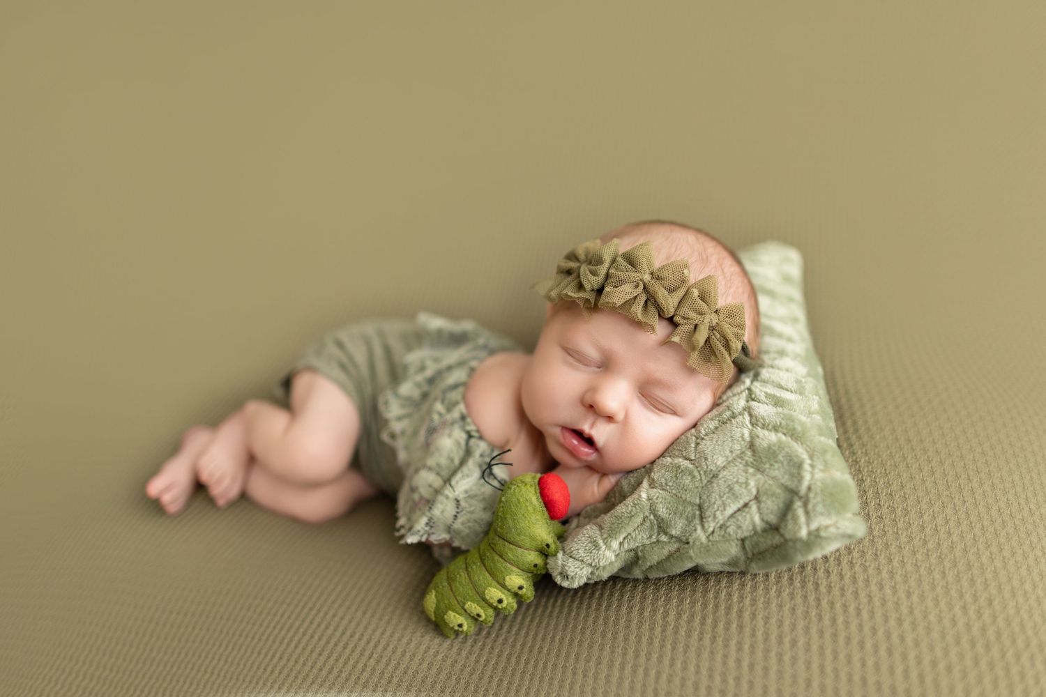 Newborn baby sleeping peacefully in green romper with floral headband, cuddling a toy caterpillar on a green pillow.
