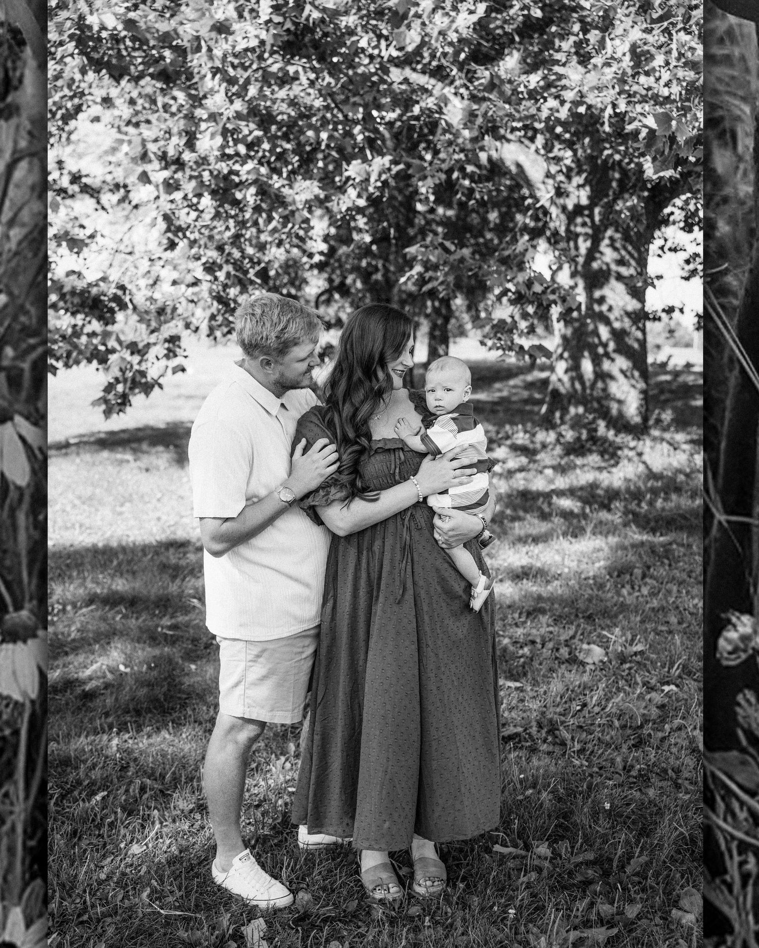 Black and white photo of a couple standing under a large tree in a sunny park, woman in dress, man in casual shorts.
