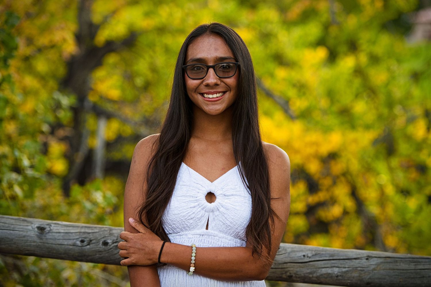 Smiling young woman with sunglasses and white top leans on wooden fence with colorful autumn foliage background.