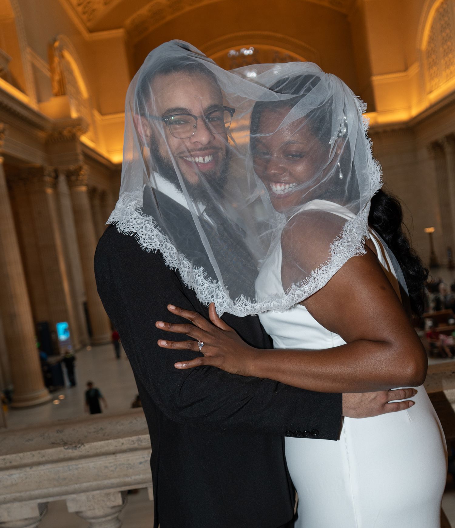 A joyful newlywed couple smiling together under a lace bridal veil in an elegant wedding venue.