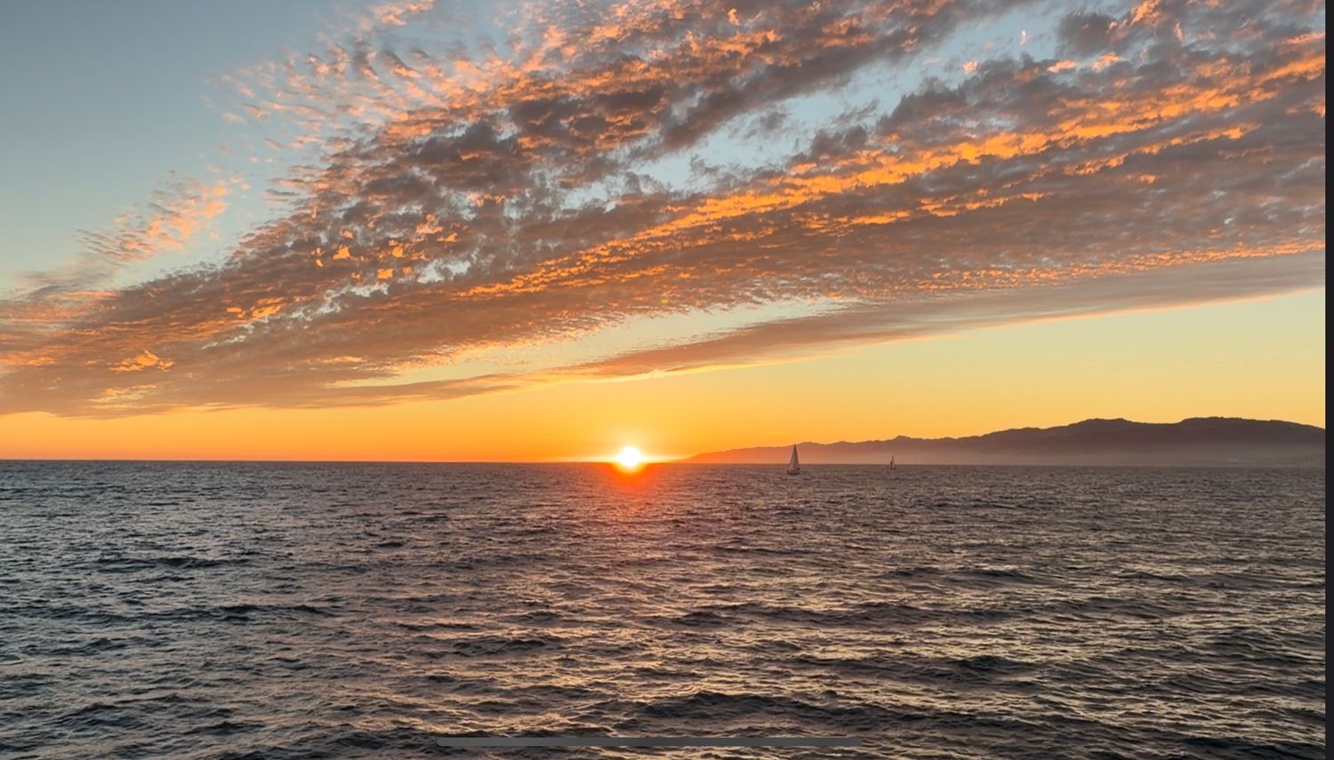 Stunning ocean sunset with vibrant orange sky, dramatic clouds, and distant mountains reflecting on calm sea waters.