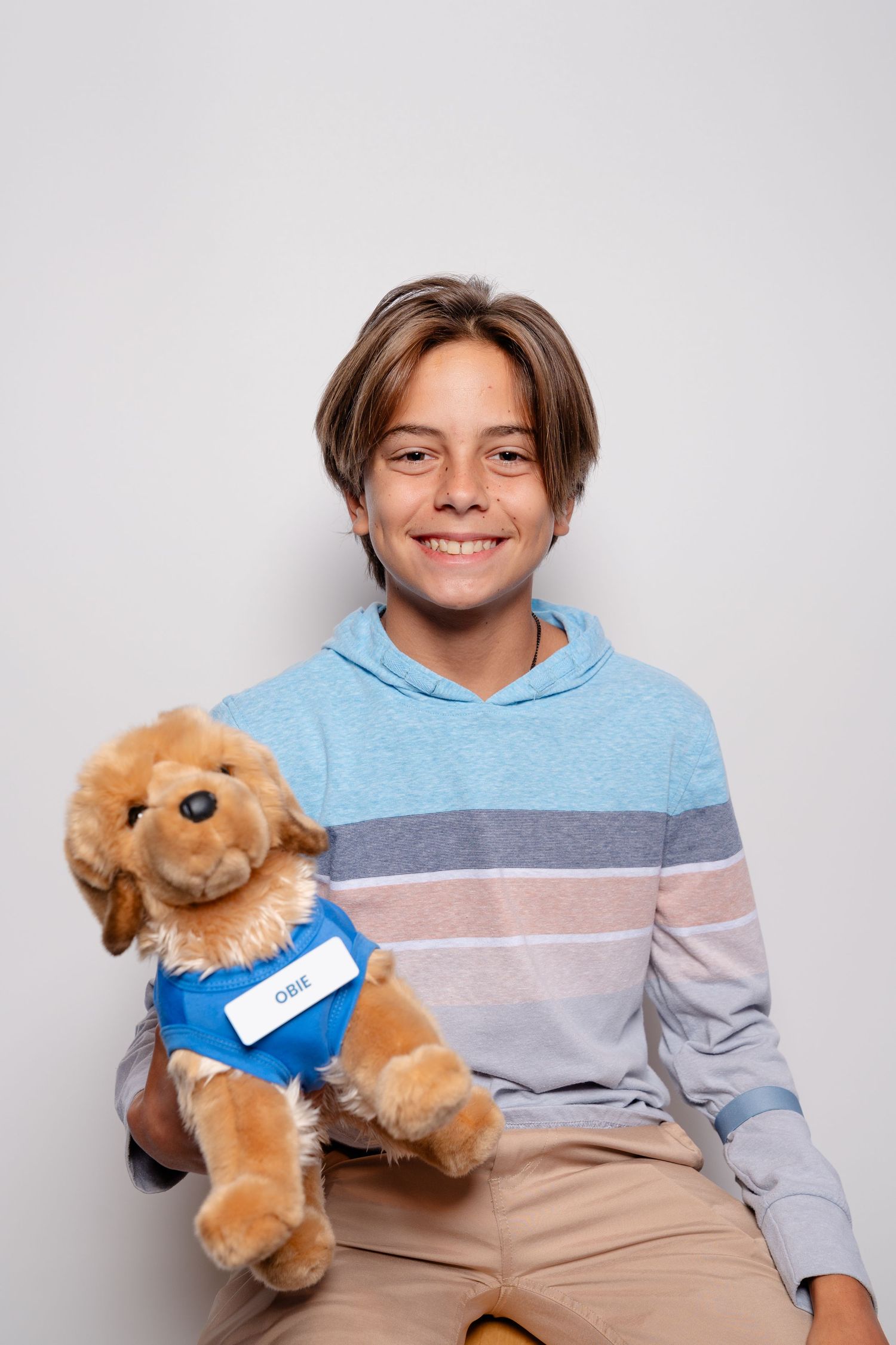 Smiling boy holding a golden stuffed animal dog toy wearing a blue vest, posing against a white background.