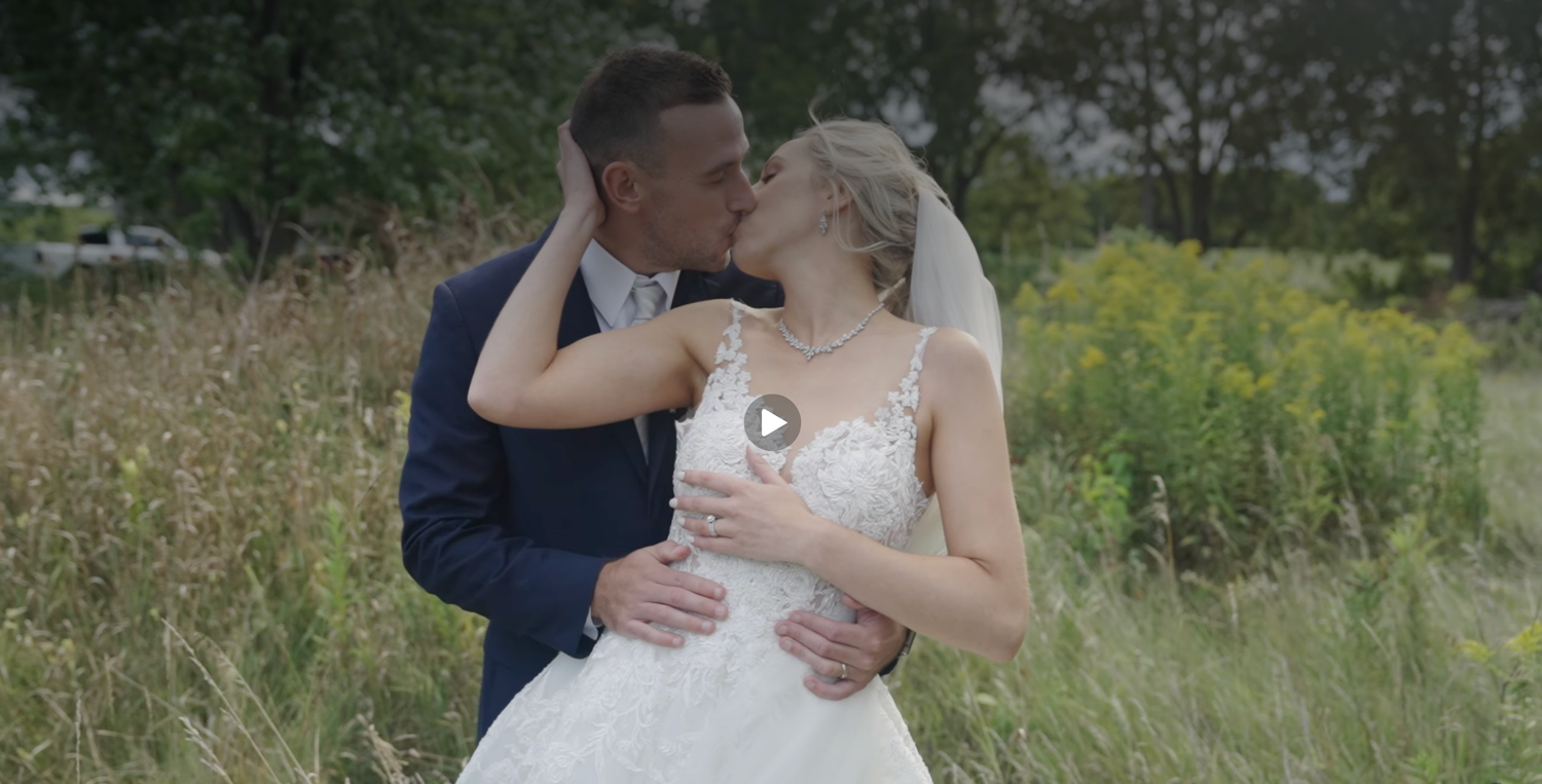 A romantic wedding couple shares a tender kiss in a sunlit field, bride in white gown and groom in navy suit.