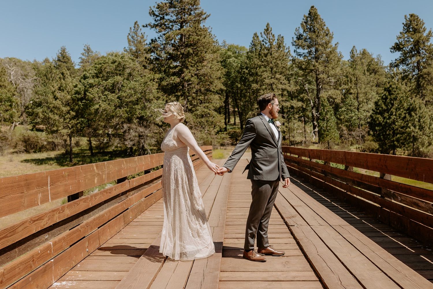 Bride and groom hold hands on a wooden bridge surrounded by pine trees on their wedding day at Lake Cuyamaca, Julian.