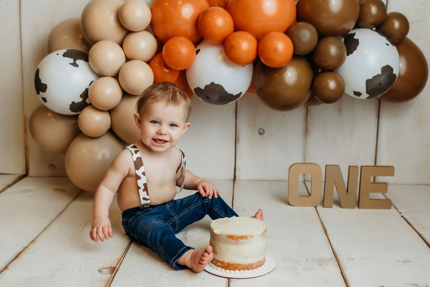 First birthday setup with brown and orange balloon arch, wooden number ONE sign, and small white cake.